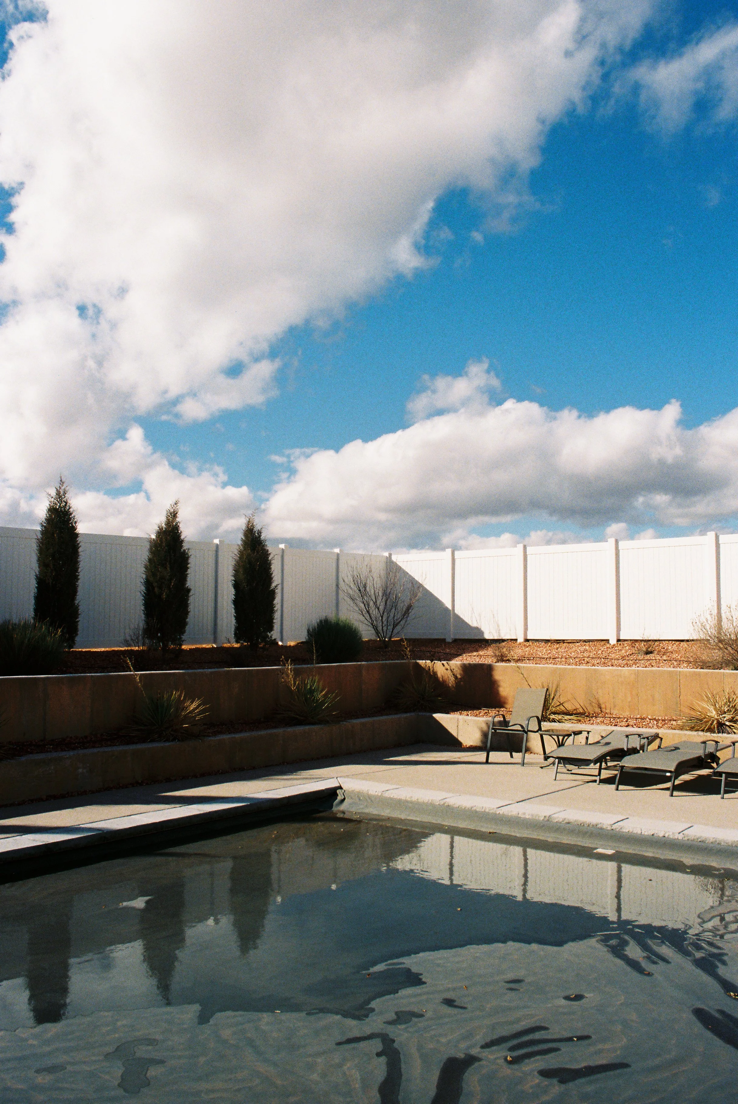 A backyard patio with a swimming pool, lounge chairs, and a white fence under a partly cloudy sky.