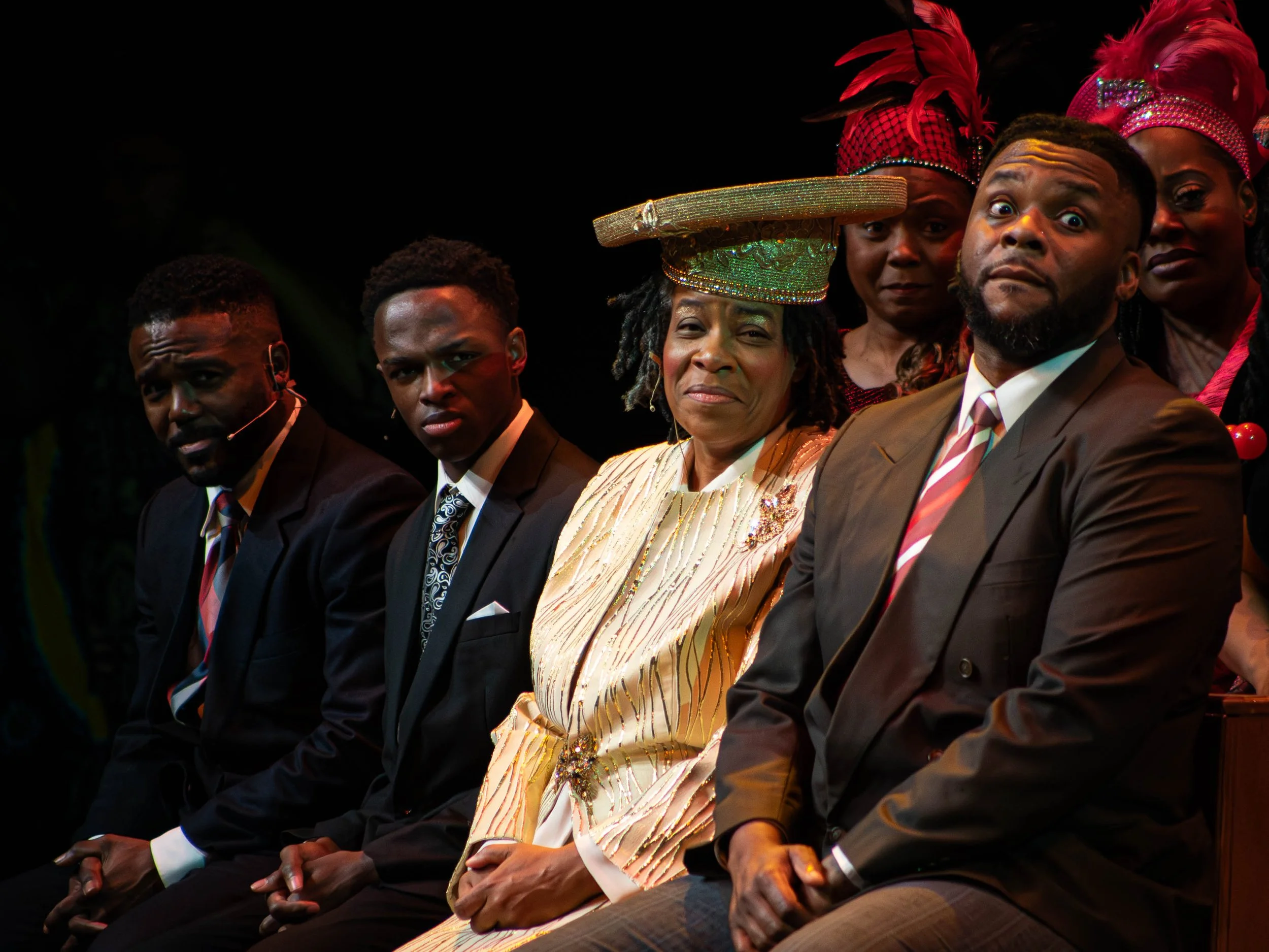 A group of people dressed formally, sitting on stage during an event. The woman in the center wears a large, decorative hat and a light-colored dress with gold accents. The men around her are in suits, and women in the background wear bright pink hea