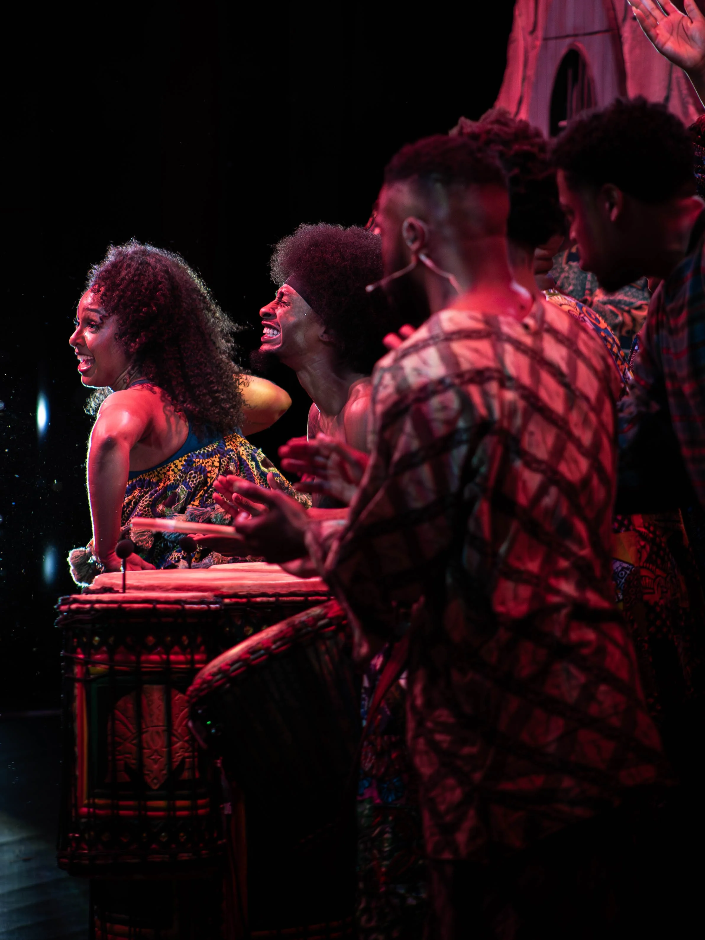 Group of singers and drummers performing on stage with joyful expressions, using traditional African drums and colorful attire.