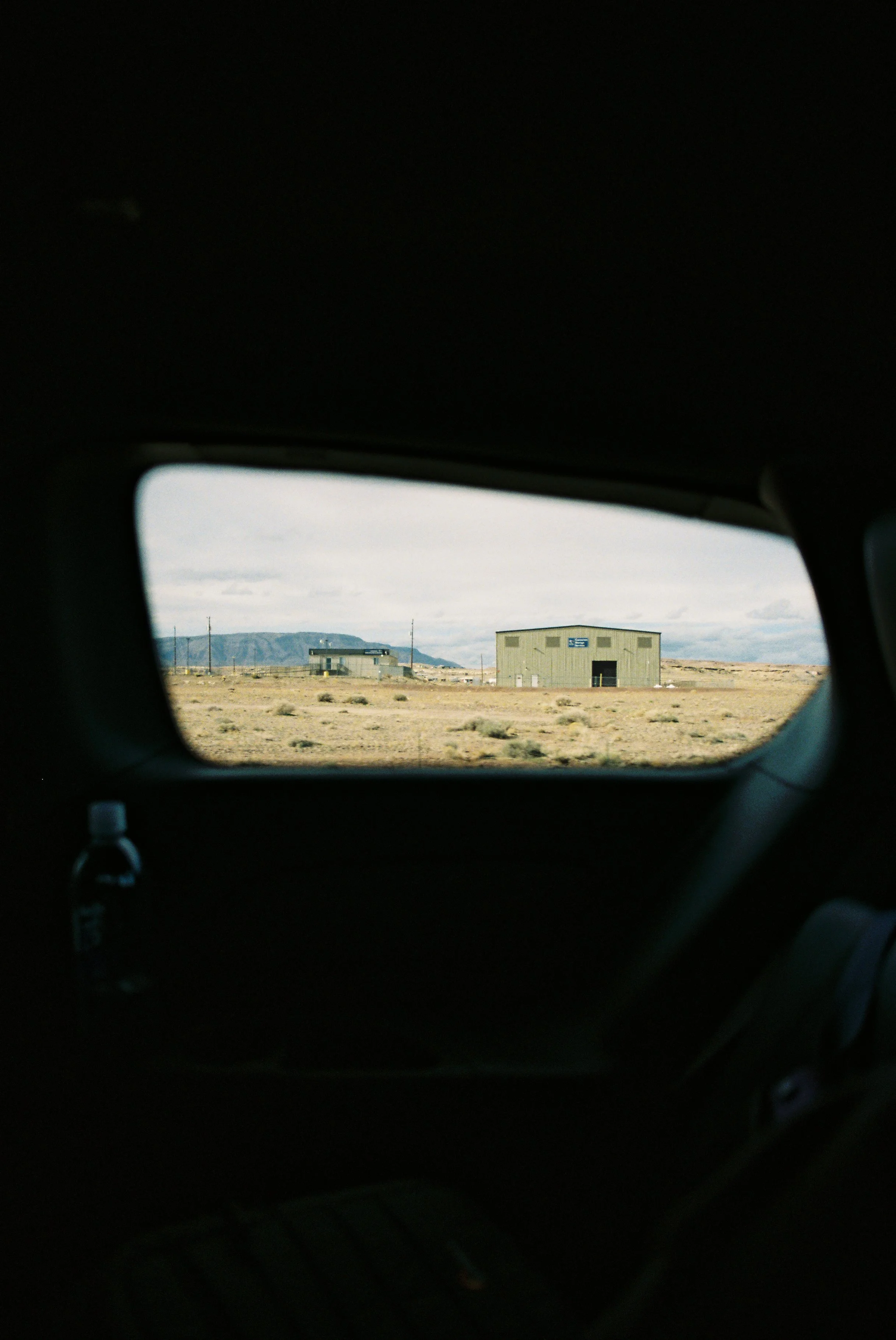 View through a car window showing an arid landscape with a small building and distant mountains in the background.