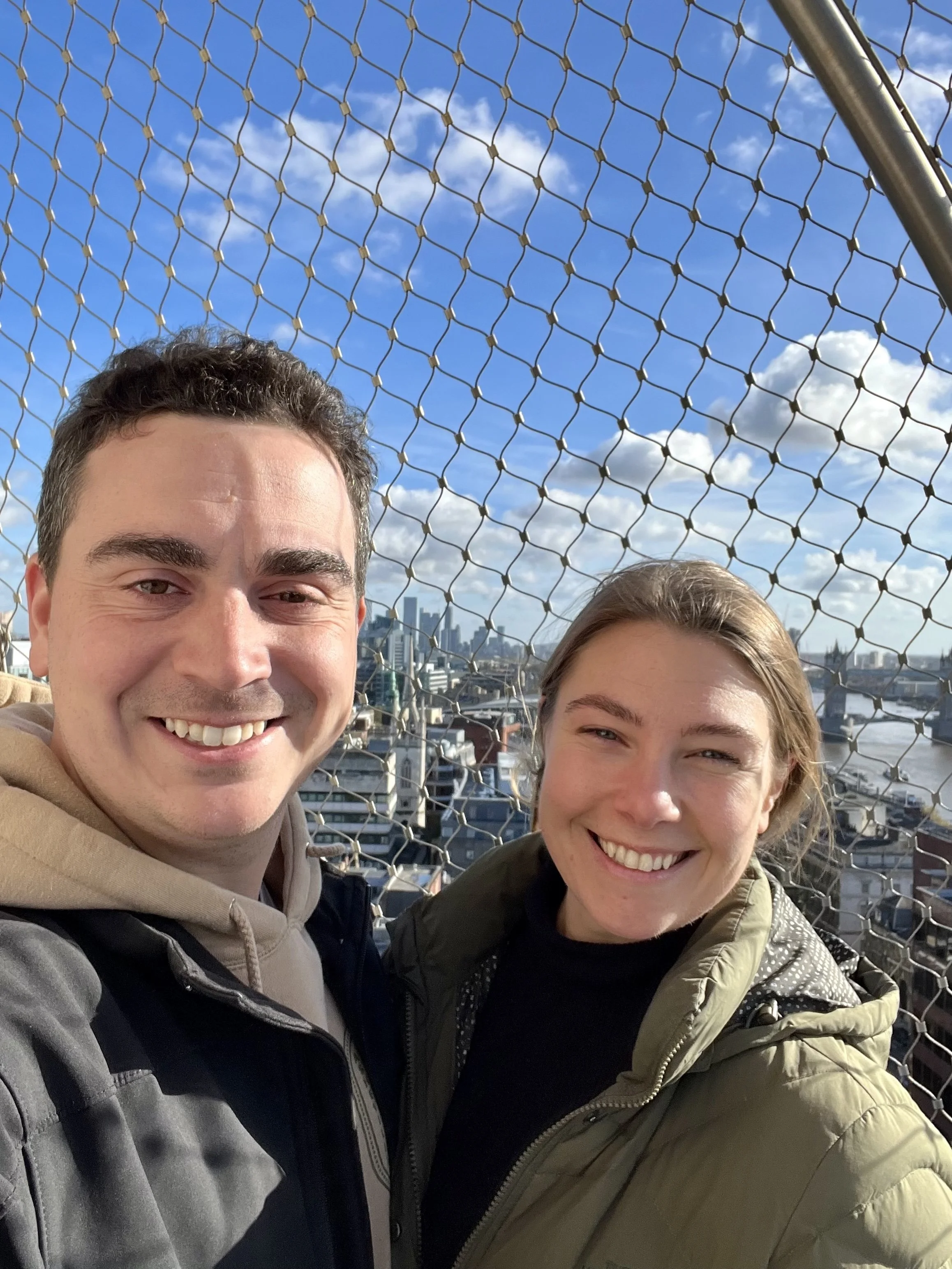 Two smiling people taking a selfie on an observation deck in London with a city skyline and a river in the background, behind a metal safety fence, during daytime with partly cloudy skies.