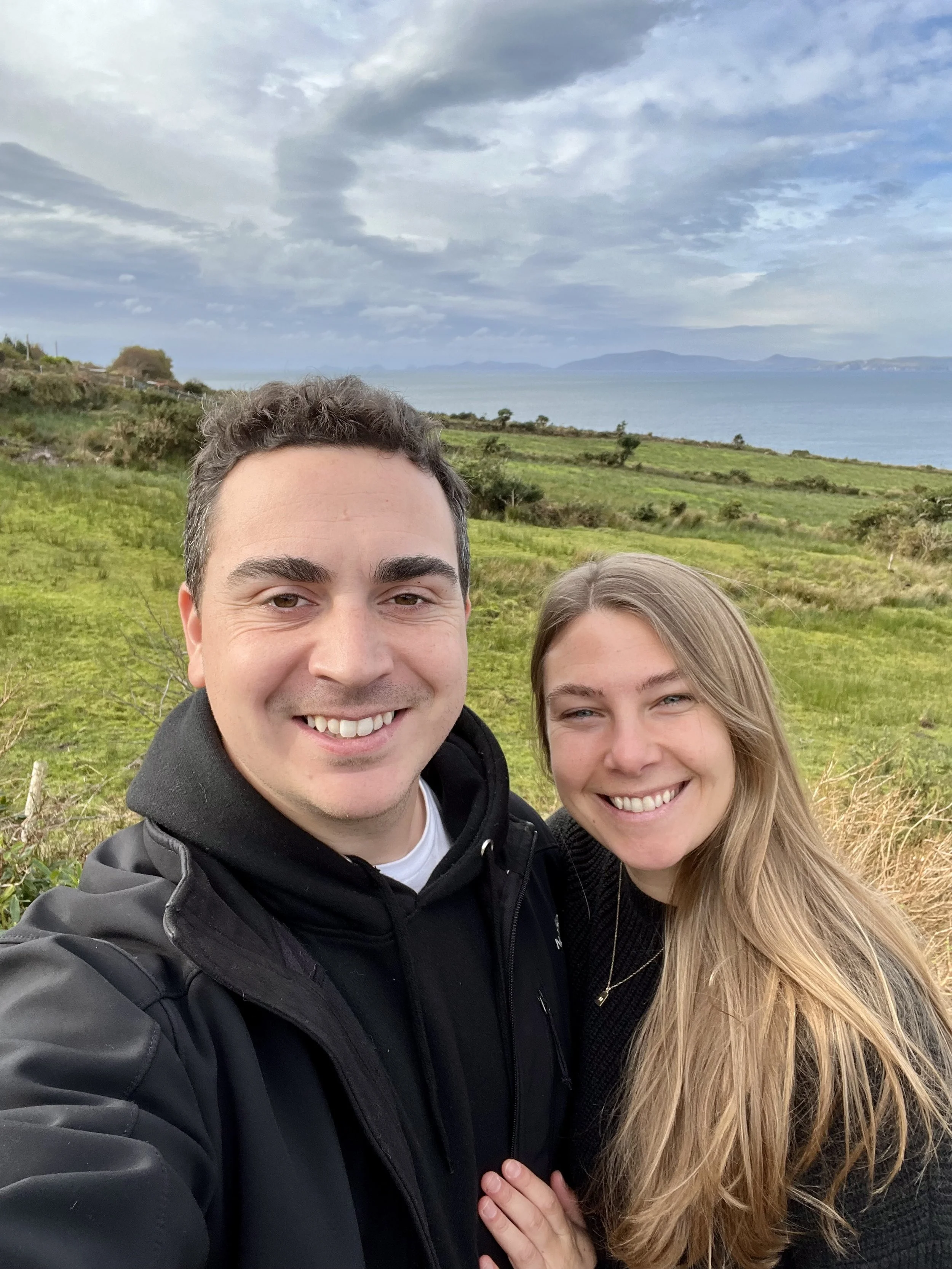 A smiling man and woman taking a selfie outdoors in Ireland with a scenic background of green fields, hills, water, and a cloudy sky.