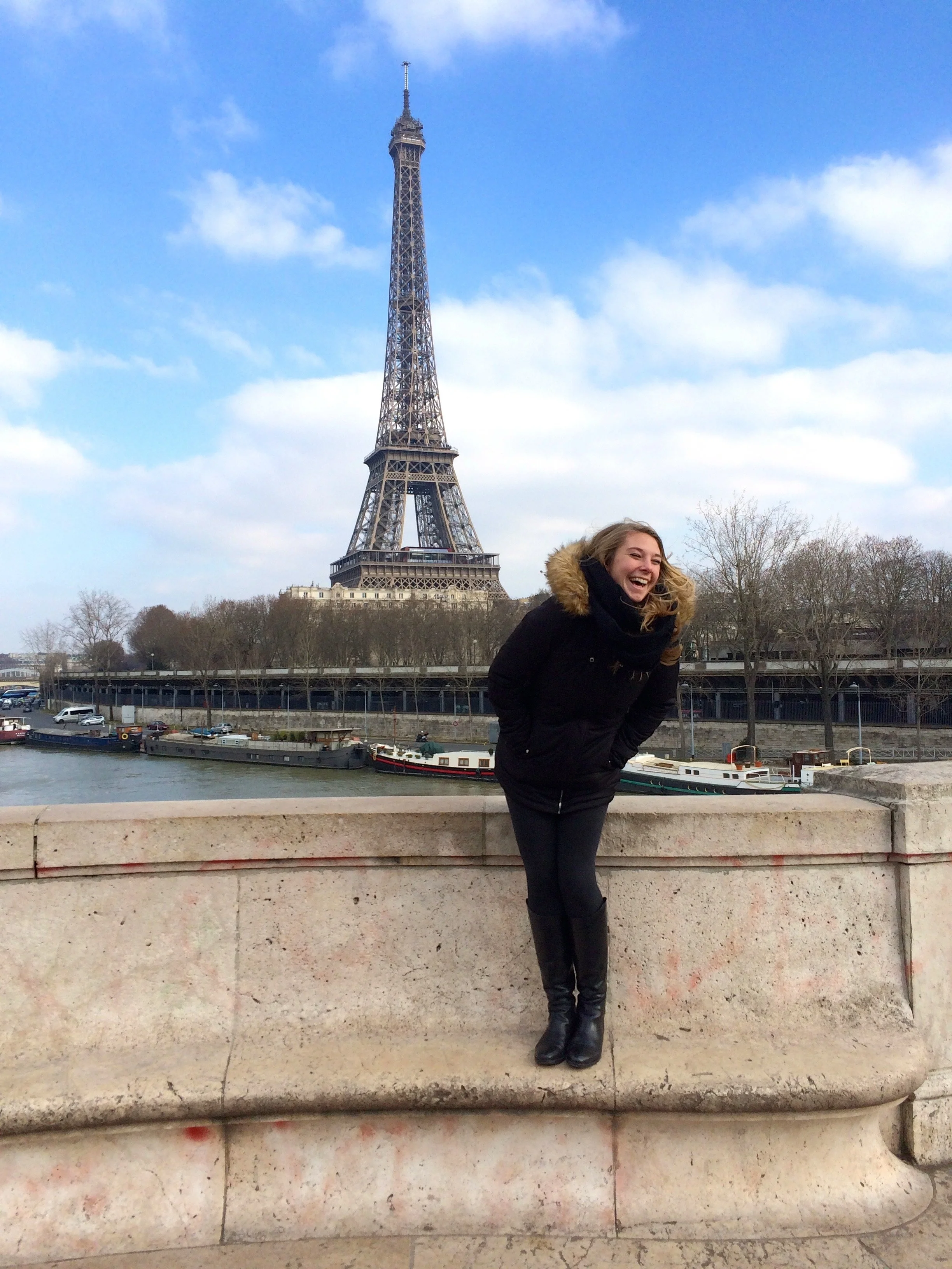A woman in a black winter coat with a fur-trimmed hood, laughing and leaning forward, standing in front of the Eiffel Tower in Paris, France, by the river with boats in the background.