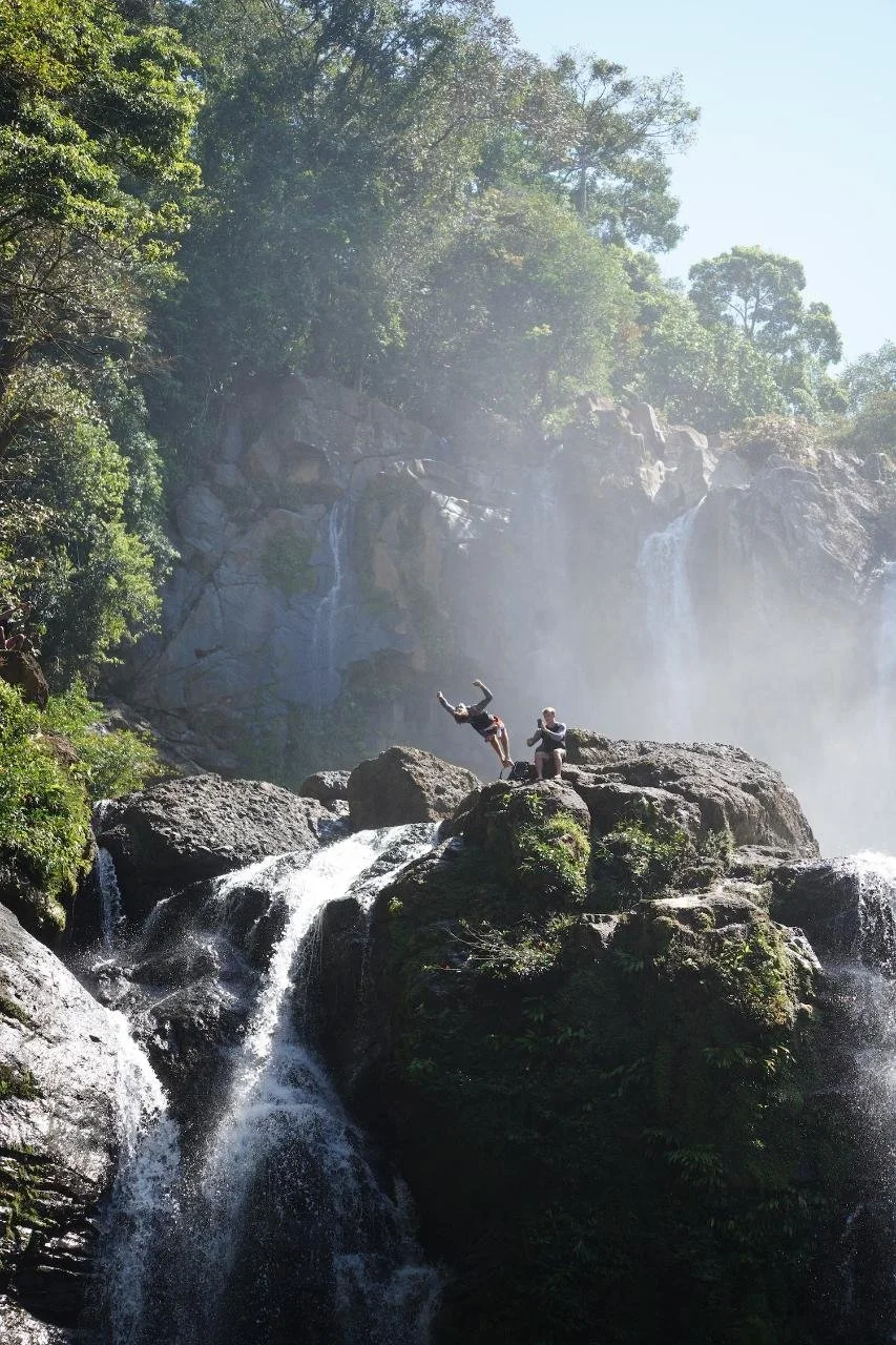People cliff jumping in costa rica and relaxing on large rocks at the base of Cataratas Nauyaca waterfall, surrounded by lush greenery and tall trees in the jungle .