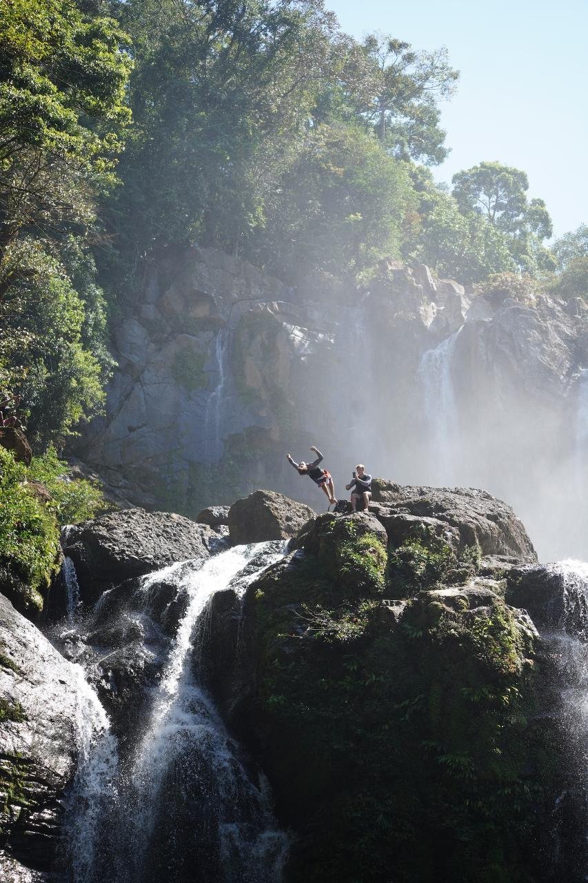 people on jumping off rocks by a waterfall in a lush green forest, one appears to be diving or jumping into the water while the other sits on the rocks.