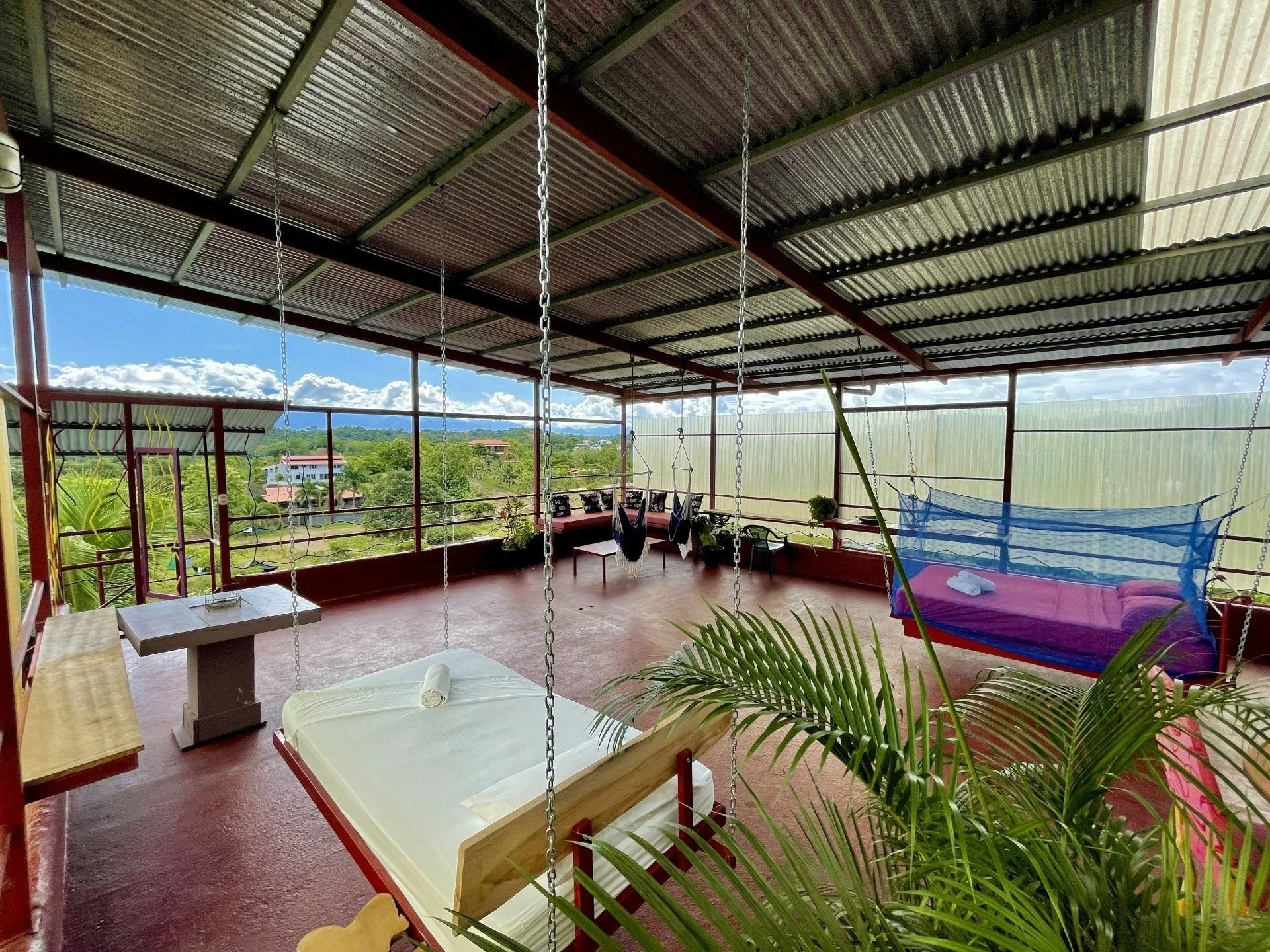 A covered outdoor patio area with swings, a bed, hanging chairs, and outdoor furniture, with a scenic view of green hills and houses in the distance.
