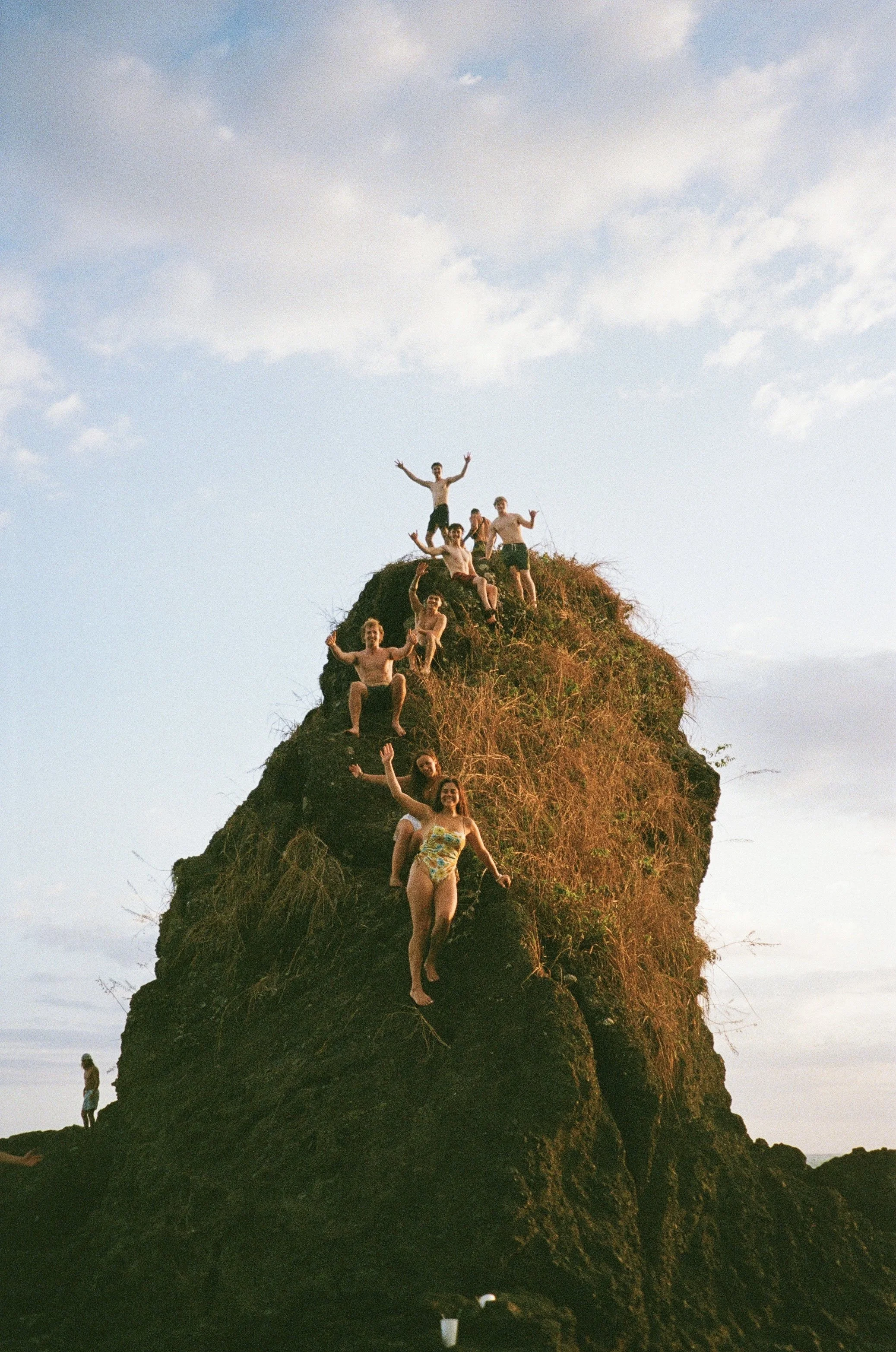 A group of people standing and sitting on top of a large rocky formation at sunset, with one person standing at the bottom. The sky is partly cloudy.