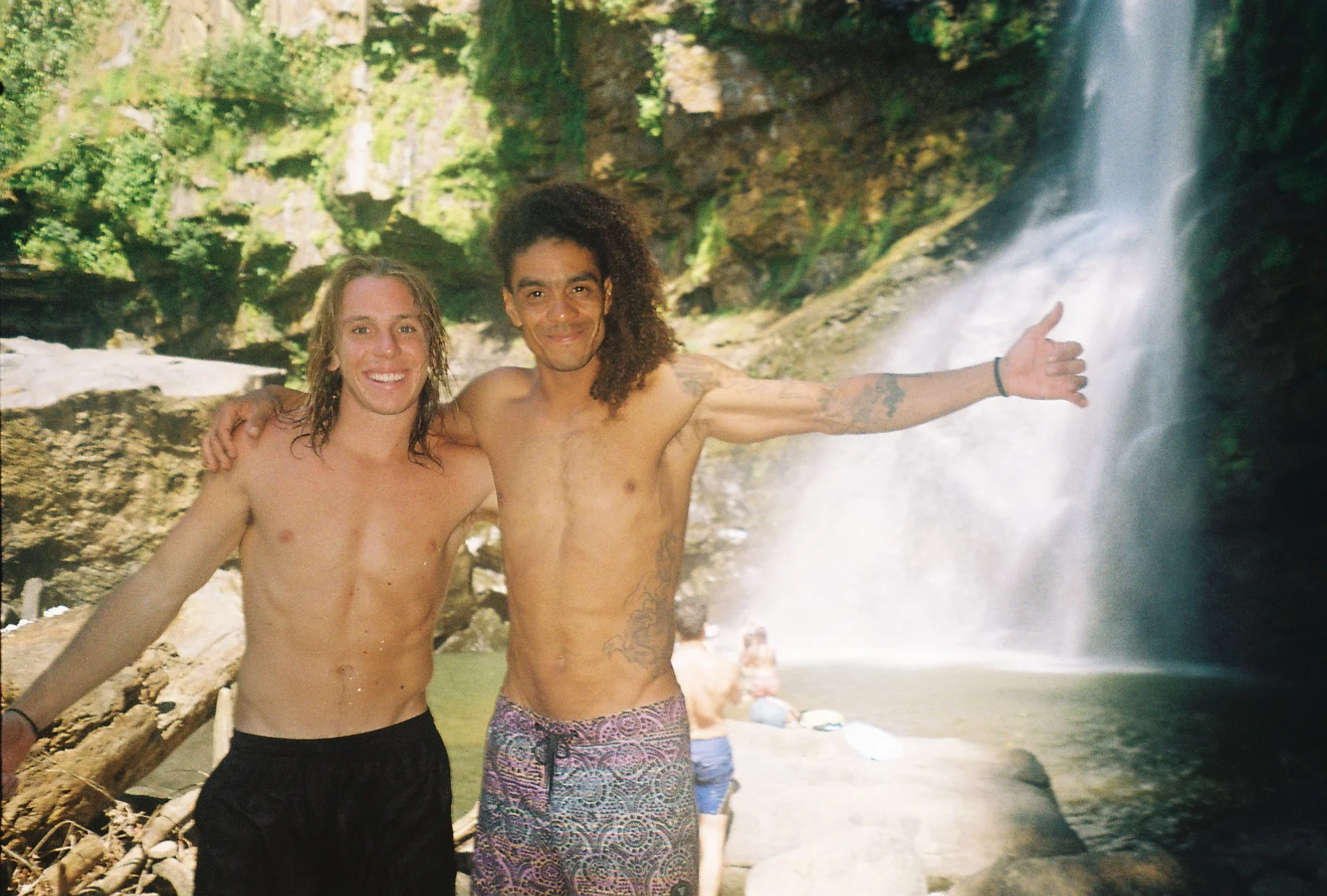 Two shirtless men with long hair standing by a waterfall in a forest, smiling and posing for the photo.