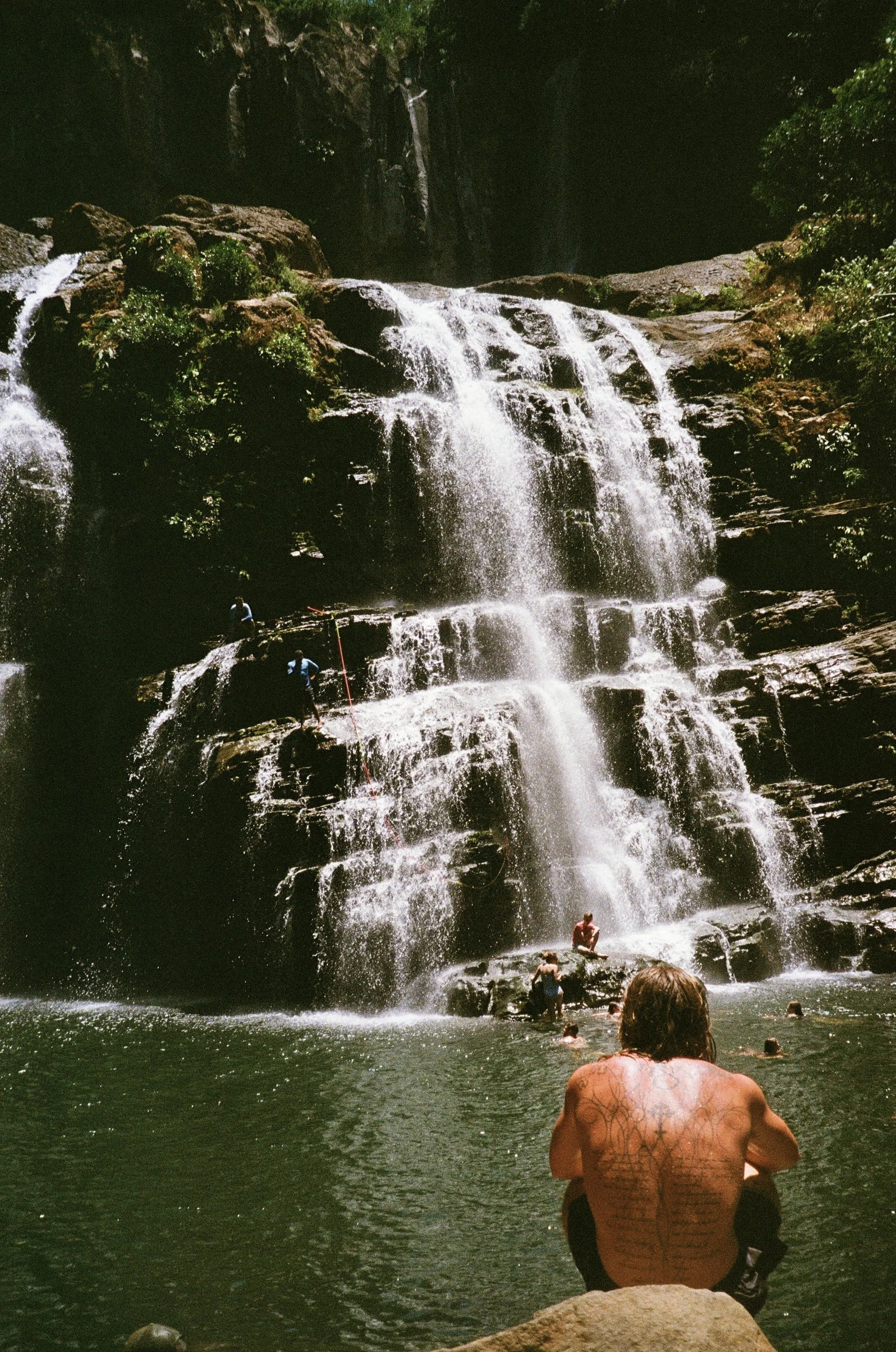 A multi-tiered waterfall cascading down rocks in a natural setting, with people swimming and sitting in the water at the base of the waterfall.