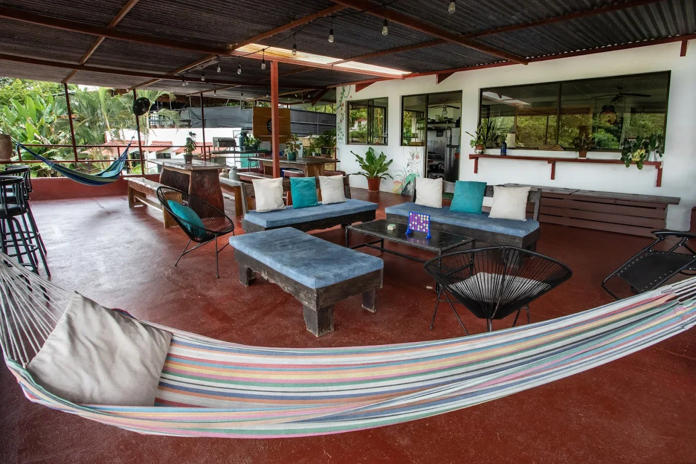 Covered outdoor patio with colorful hammock, cushioned benches, black metal chairs, potted plants, and a view of greenery outside.