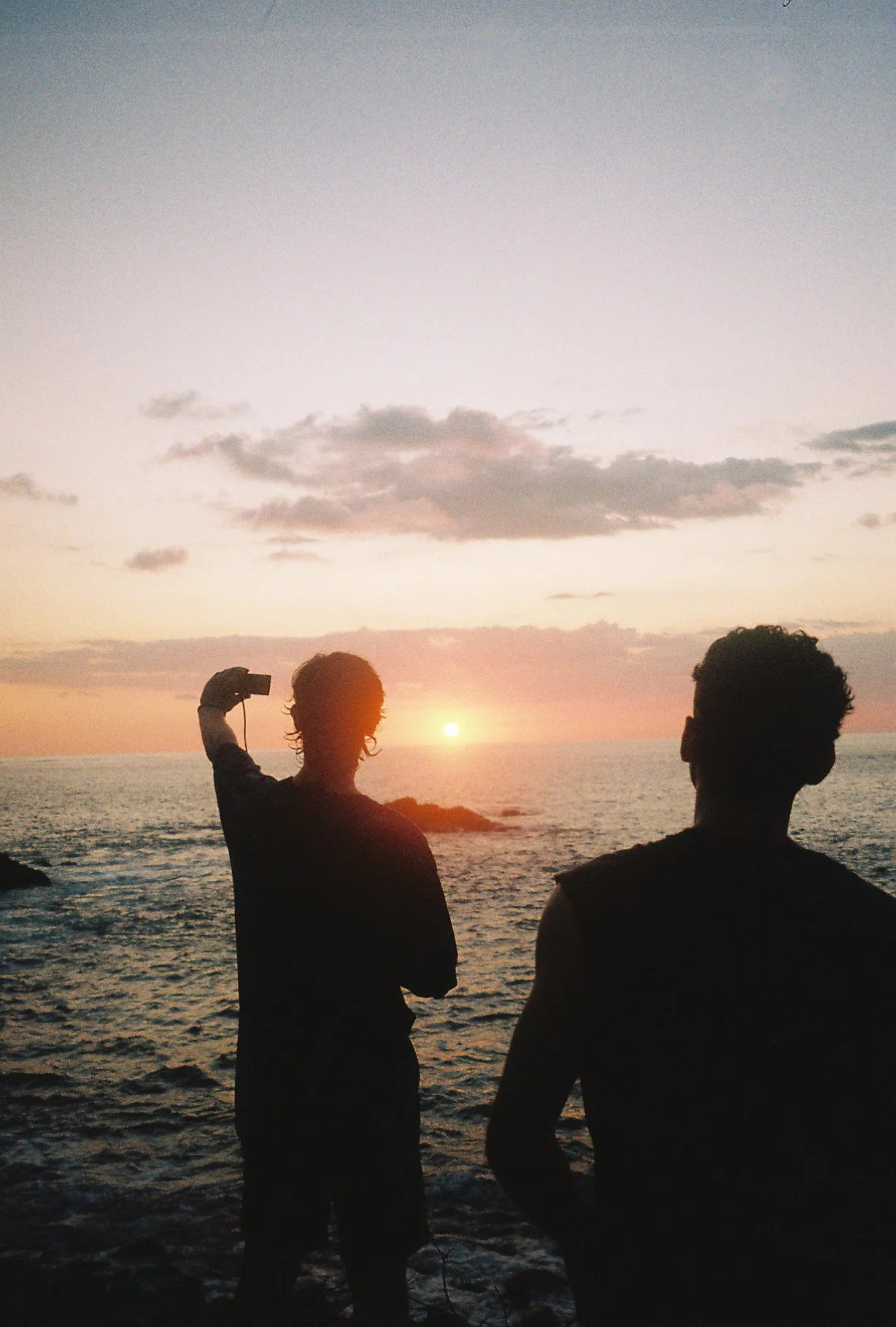 Silhouettes of two people near the ocean during sunset, one person taking a photo , colorful sky with scattered clouds.