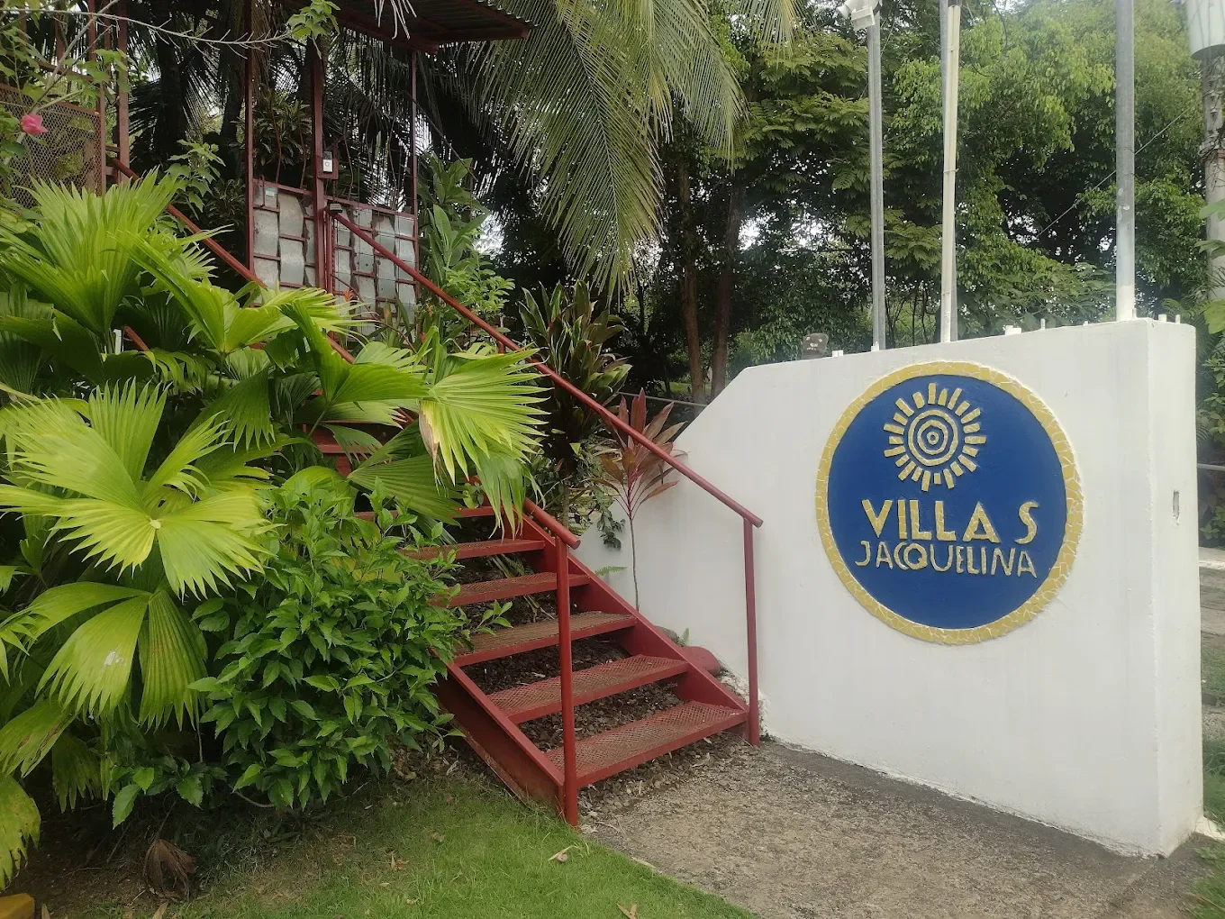 Entrance of Villas JacQUELINA with a blue and yellow sign, a red staircase, and lush tropical plants surrounding the area.