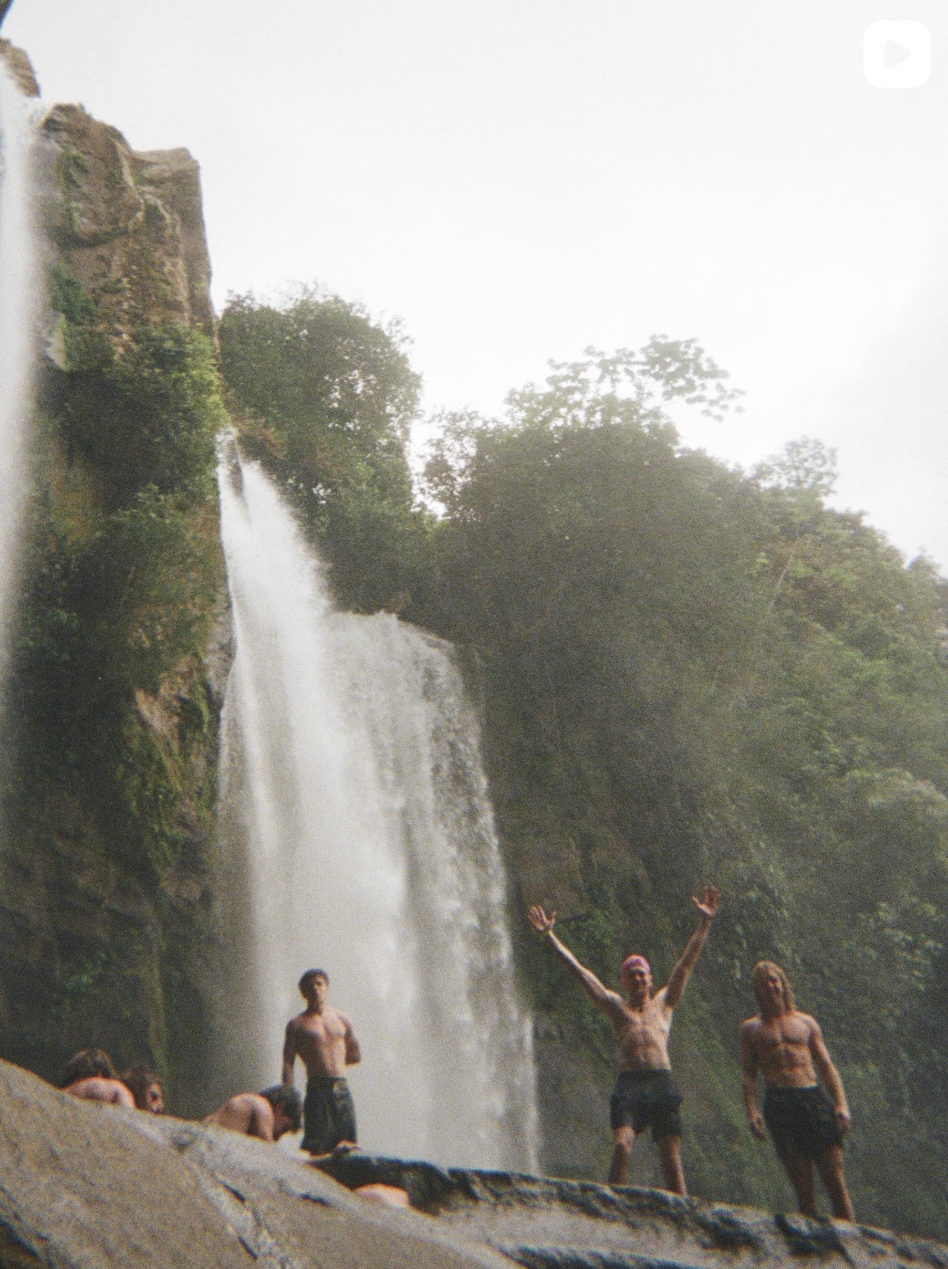 Young people and cliff jumpers swimming and standing near a waterfall in a lush, green forest