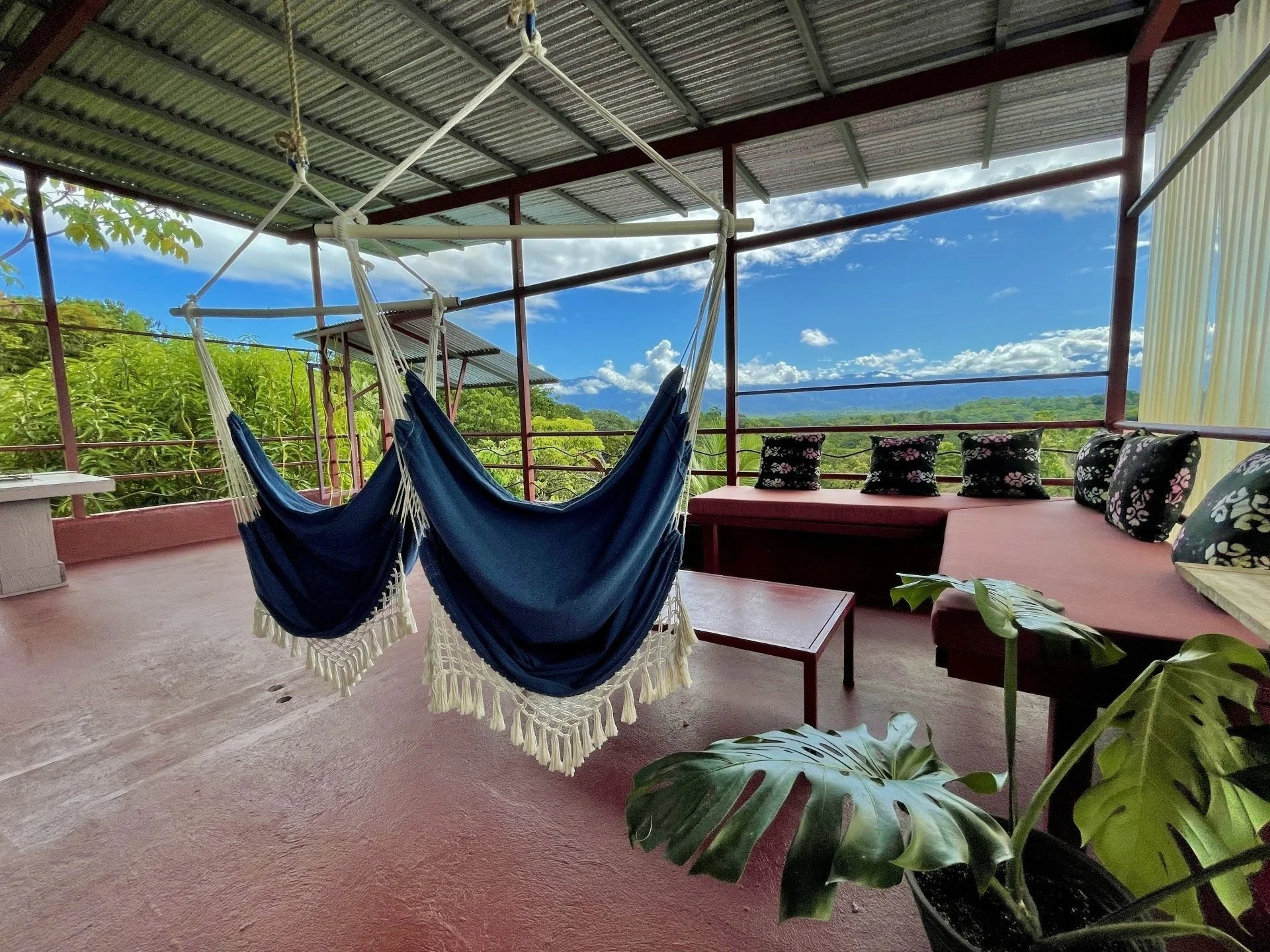 A covered outdoor patio with a blue hammock hanging from the ceiling, a pink cushioned bench with black and pink floral pillows, and potted plants, with lush green trees and mountains in the background under a partly cloudy sky.
