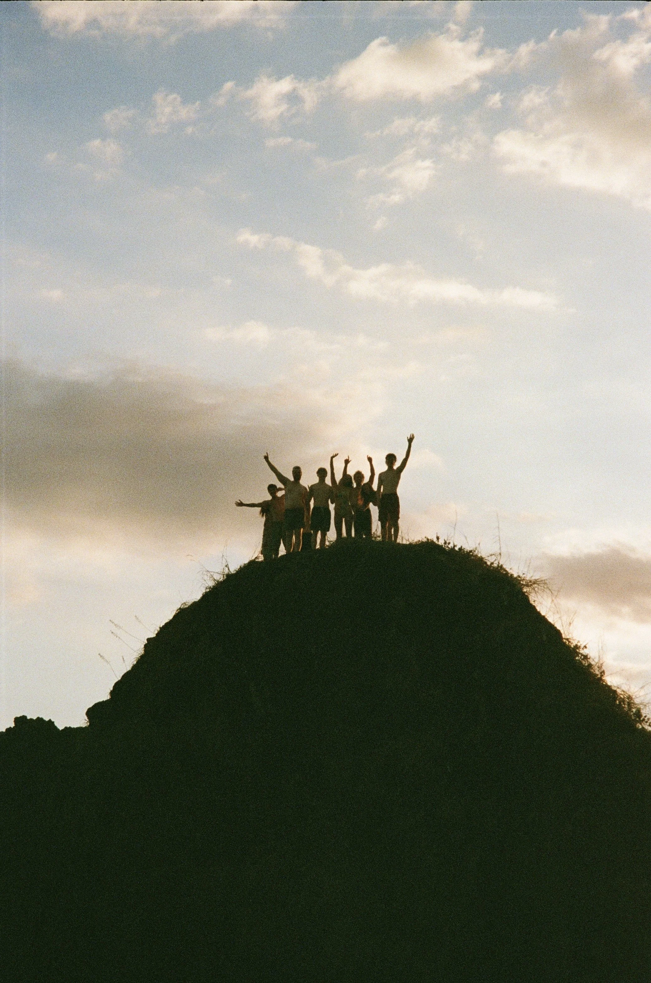 Silhouette of seven people standing on a hilltop with their arms raised, against a sky with scattered clouds during sunset or sunrise.