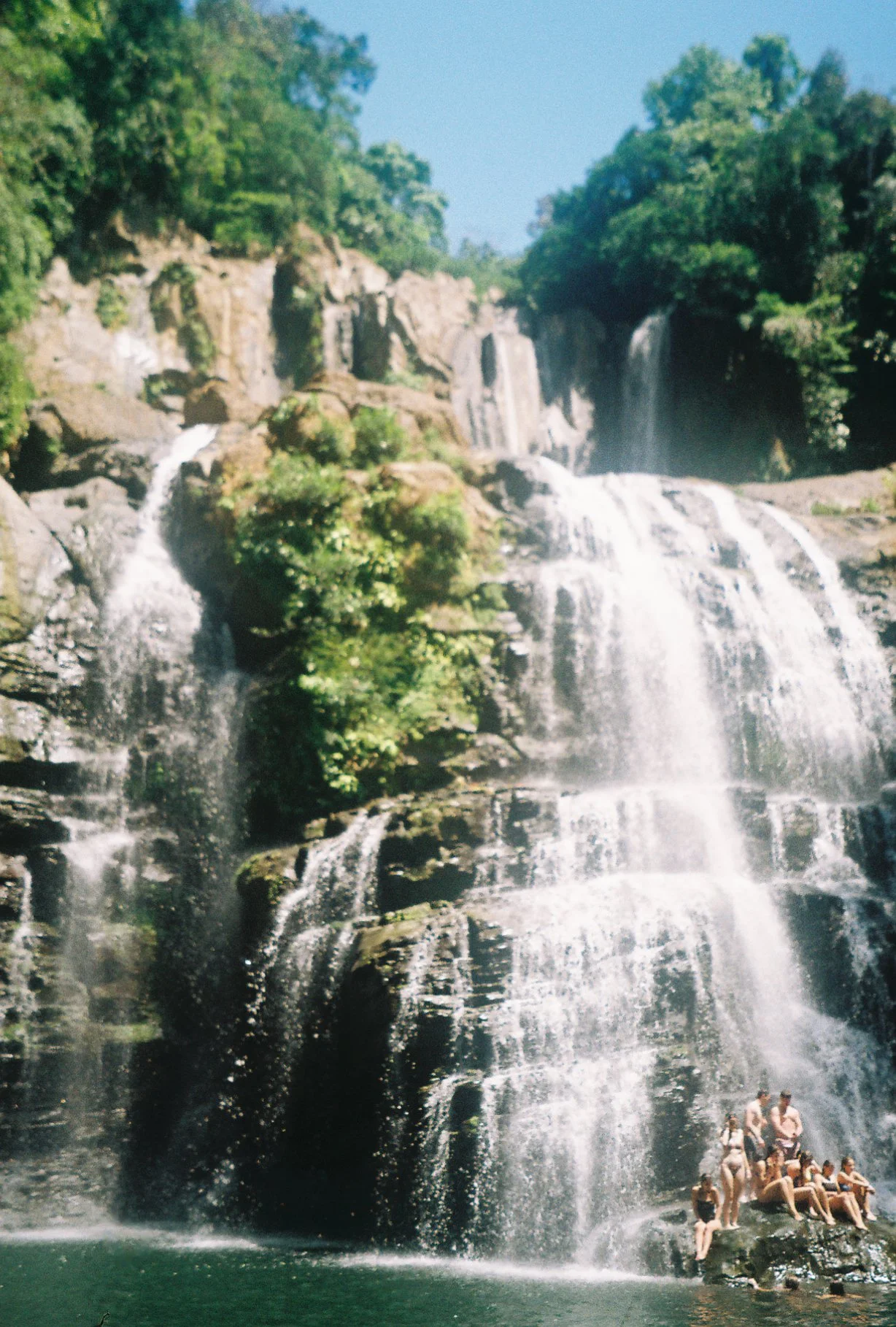Waterfall with multiple streams cascading over rocks and lush green trees surrounding it, with people sitting on rocks at the base.