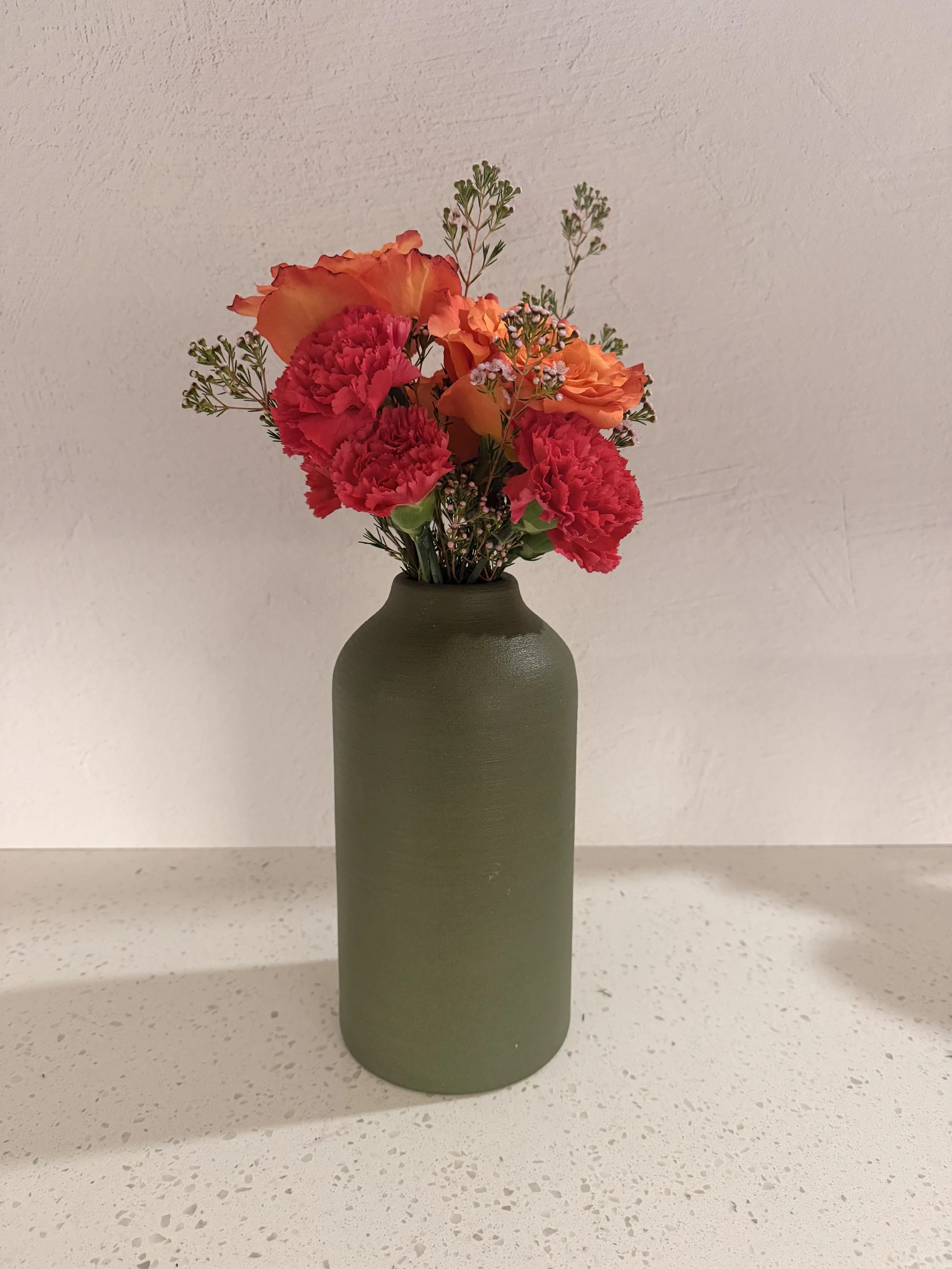 A green ceramic vase with a textured surface holding a bouquet of pink carnations, orange roses, and small white flowers, placed on a white speckled countertop against a white wall.