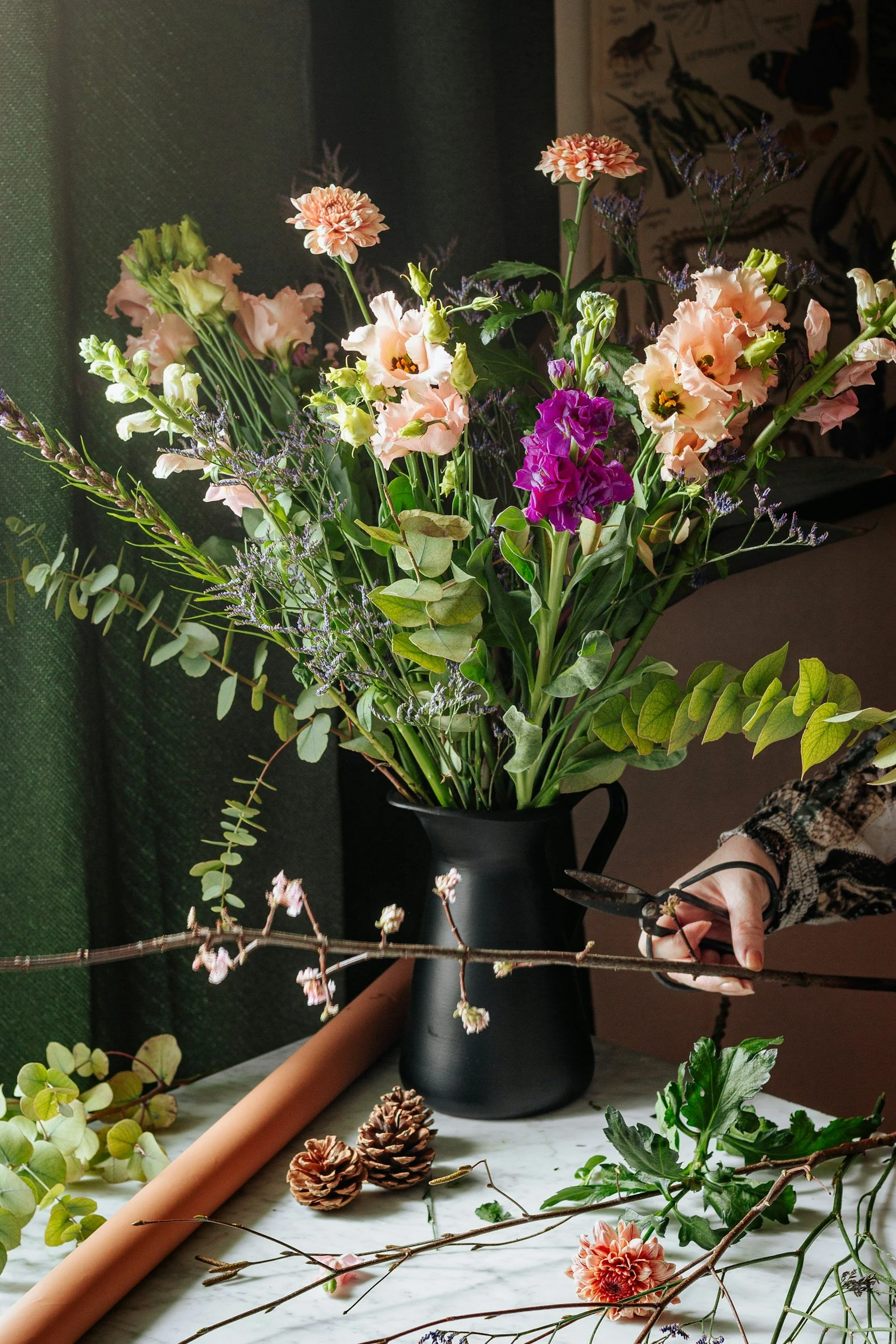 A person arranging a bouquet of mixed flowers including pink, purple, and peach blooms in a black vase on a marble surface, with floral supplies and greenery around.