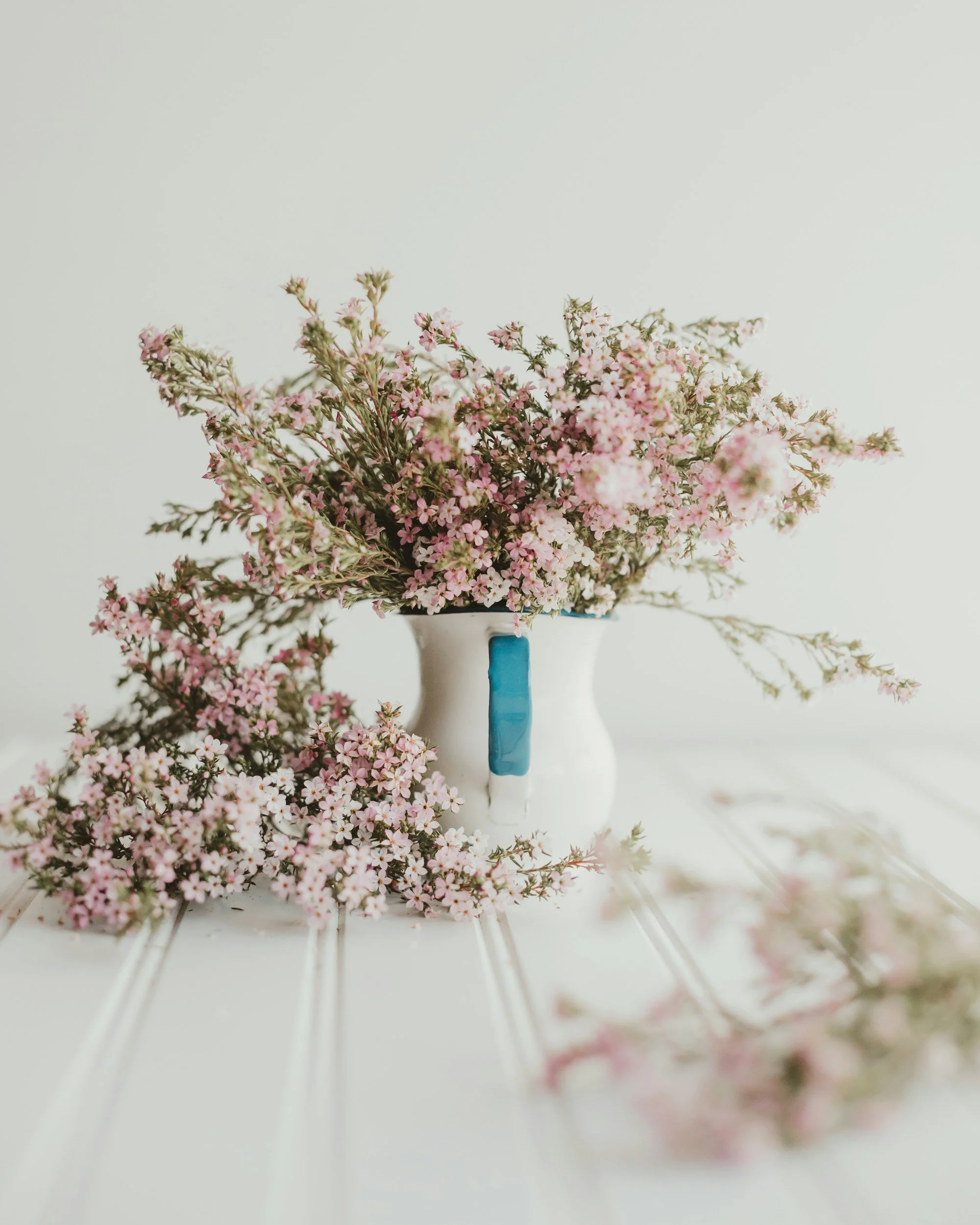 Pink and white flowers in a white vase with a blue accent, on a white surface with some flowers scattered around.