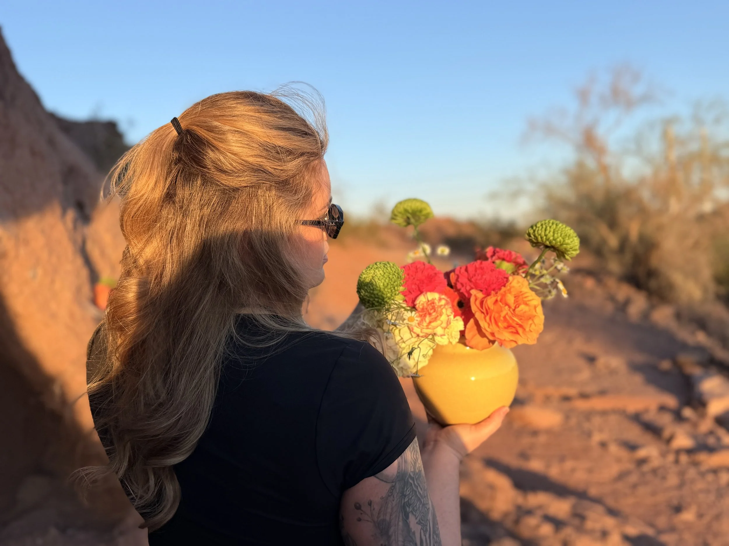 A woman with long blonde hair held in a ponytail, wearing black sunglasses, holding a yellow vase with a bouquet of colorful orange and green flowers. She is outdoors in a natural setting during sunset or sunrise, with a blue sky and blurred trees and rocks in the background.
