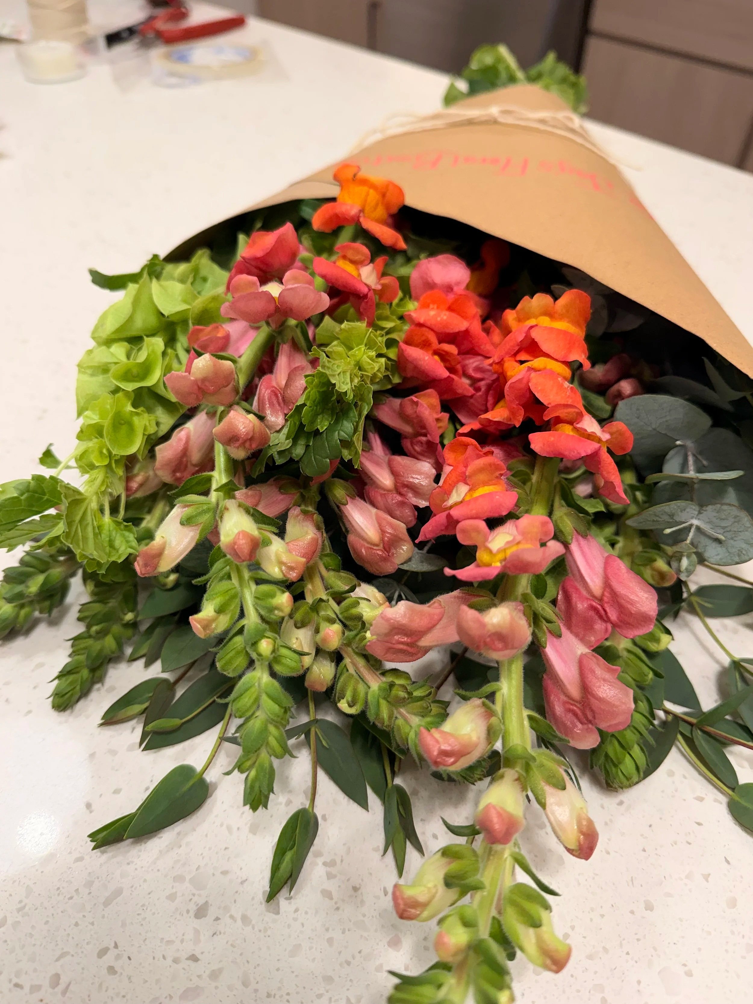 Bouquet of pink, orange, and yellow flowers wrapped in brown paper on a white surface.