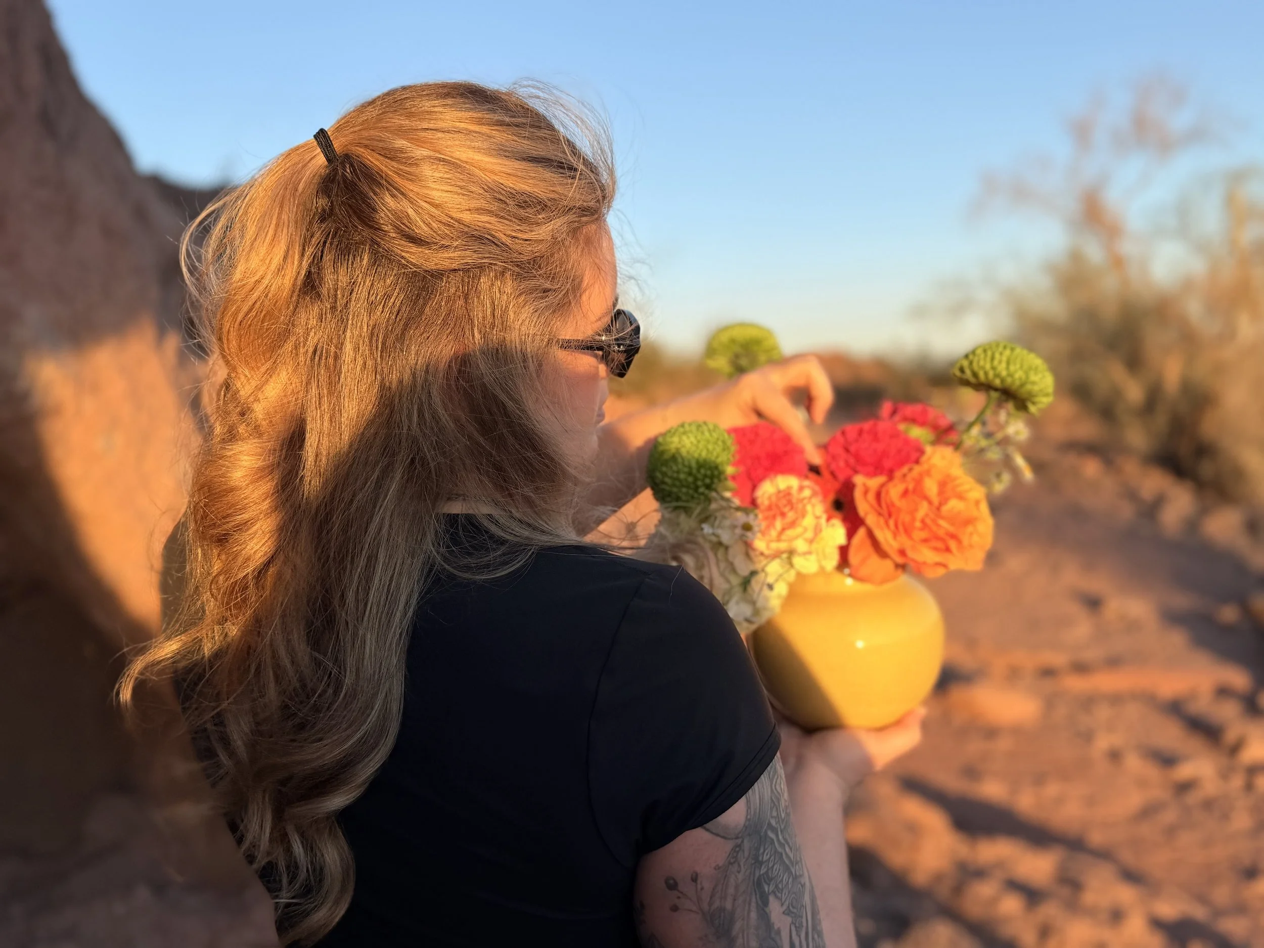 Woman with long, wavy hair holding a yellow vase filled with colorful flowers, outdoors during sunset.