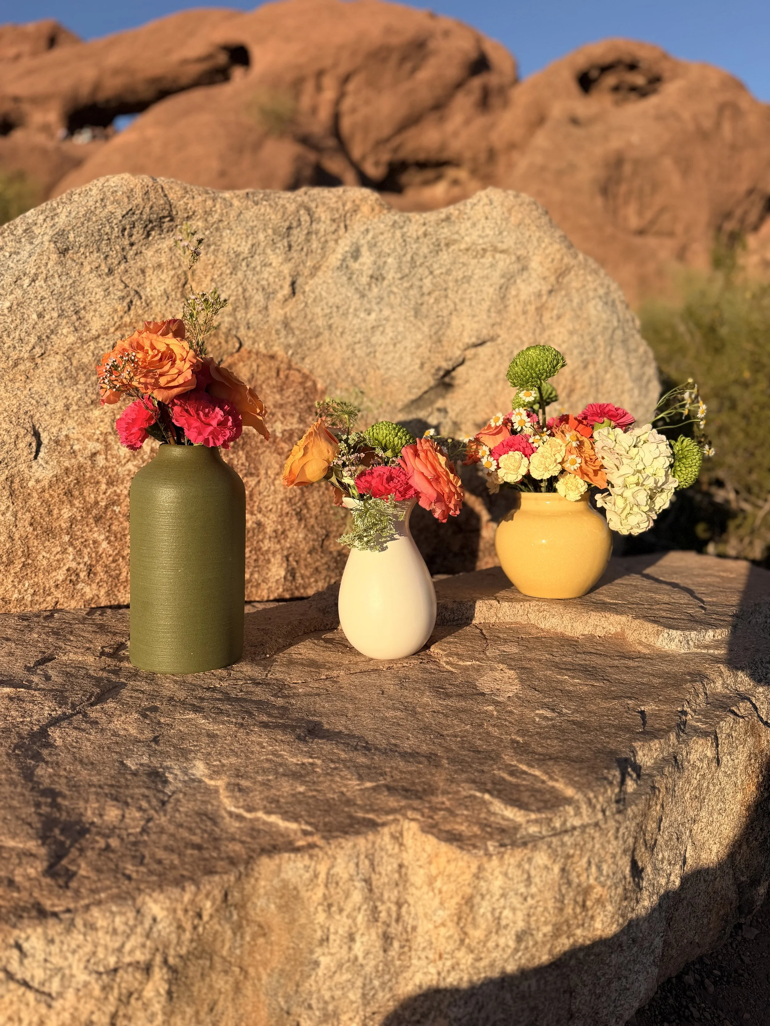 Three vases with colorful flowers on a large rock, with a desert landscape and reddish rocks in the background, taken during sunset.