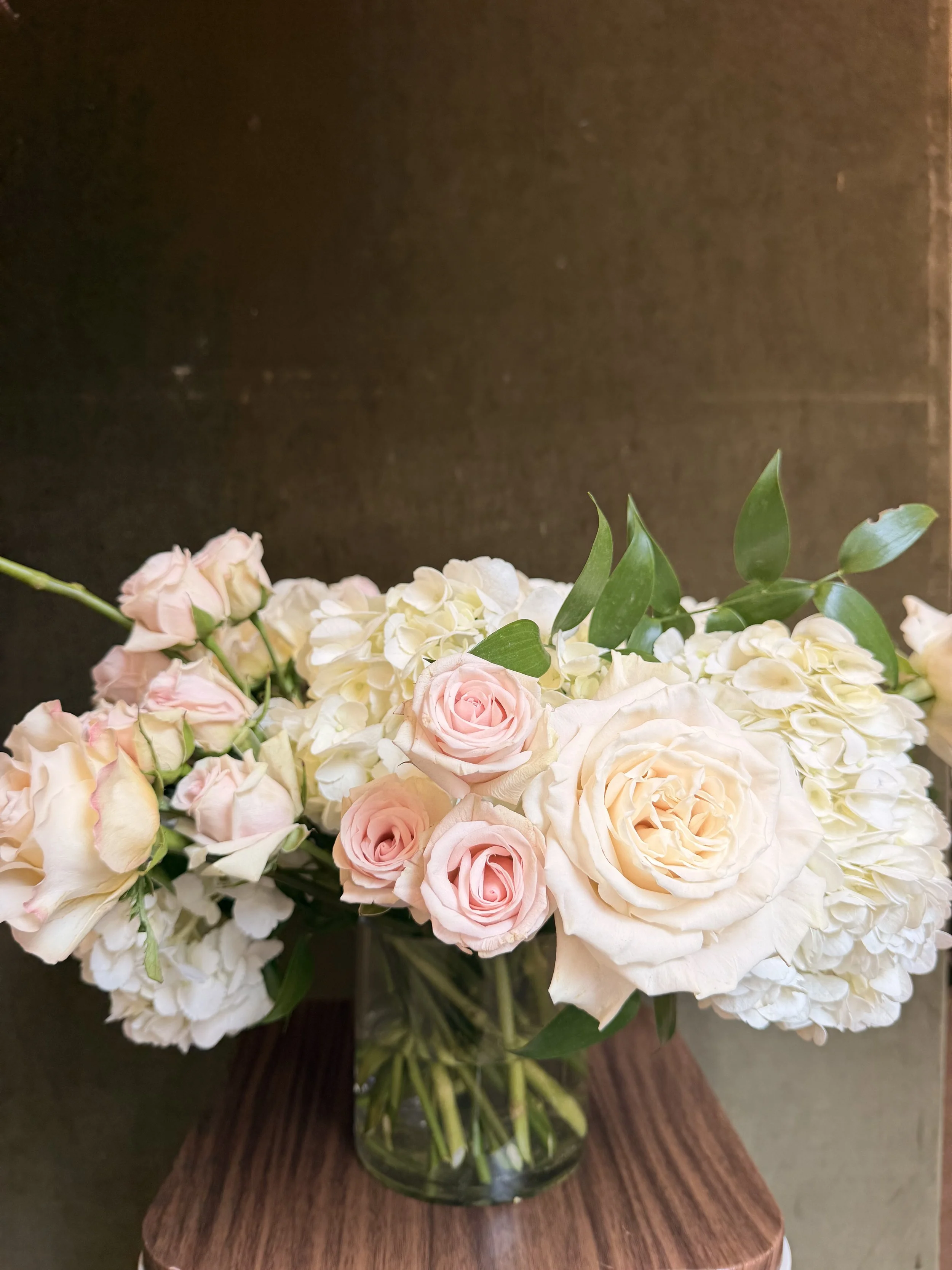 A glass vase with pink and white hydrangea flowers and green leaves on a wooden surface.