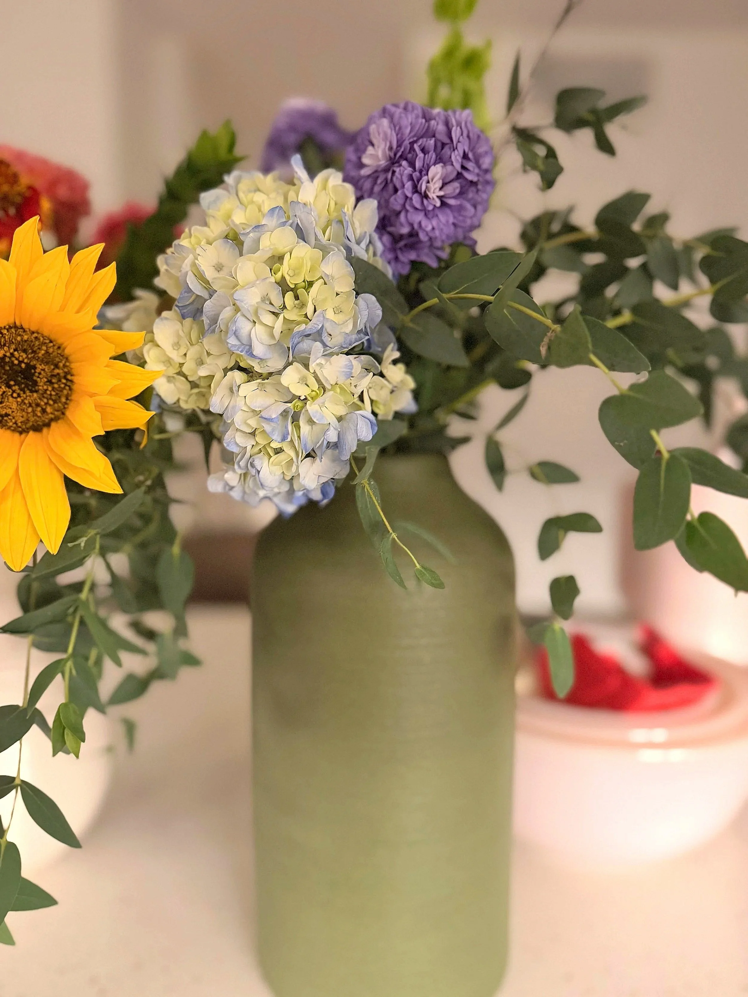 A green vase holding a bouquet of colorful flowers, including a sunflower, blue and white hydrangeas, and purple scabiosa, with green leaves.