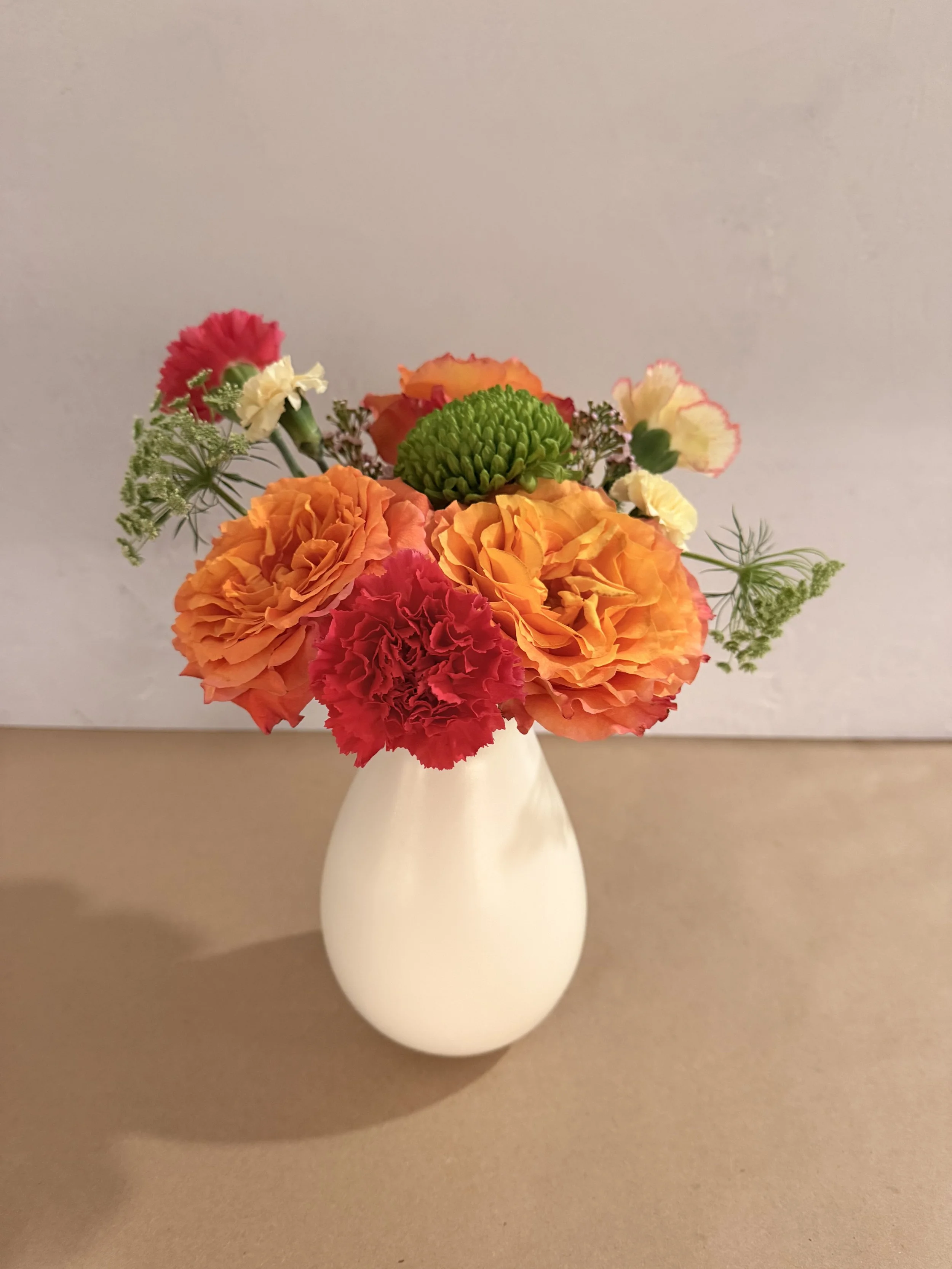 A bouquet of pink lilies, yellow daisies, and other flowers in a clear textured glass vase, with shadows cast on a white surface and wall. A smaller potted green plant and decorative candles are also on the surface.