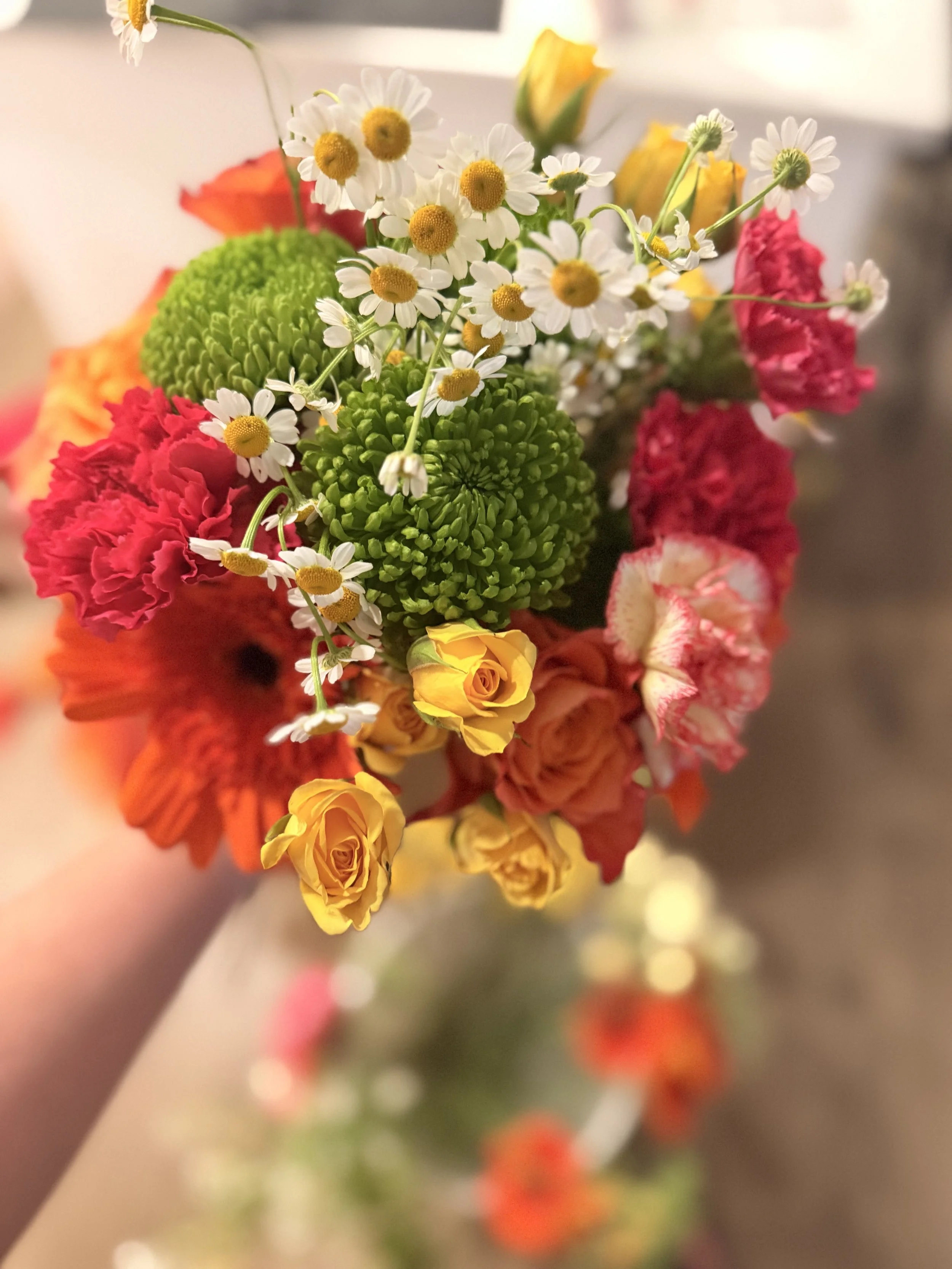 Colorful bouquet of flowers including daisies, green chrysanthemums, yellow roses, orange gerbera daisies, and pink and white flowers with a blurred background.