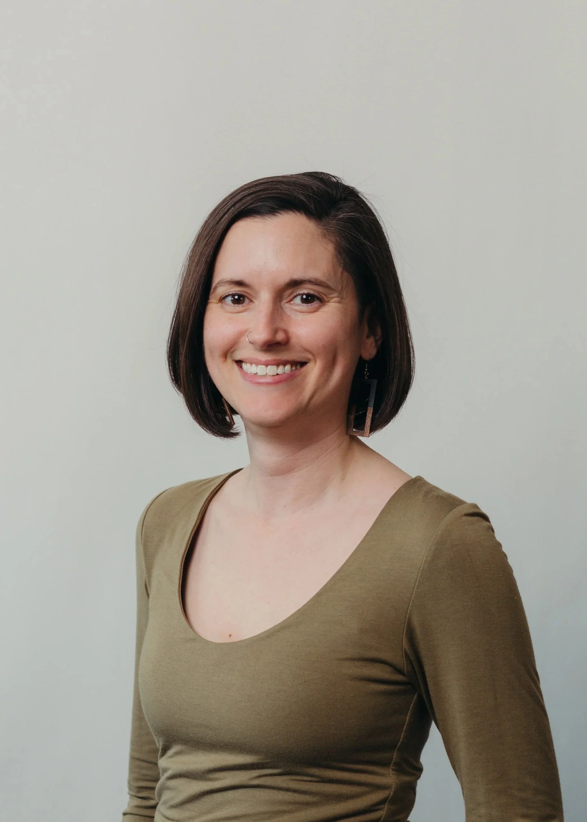 A woman with short dark hair and a nose piercing, smiling and looking at the camera, wearing a brown V-neck top, against a plain light grey background.