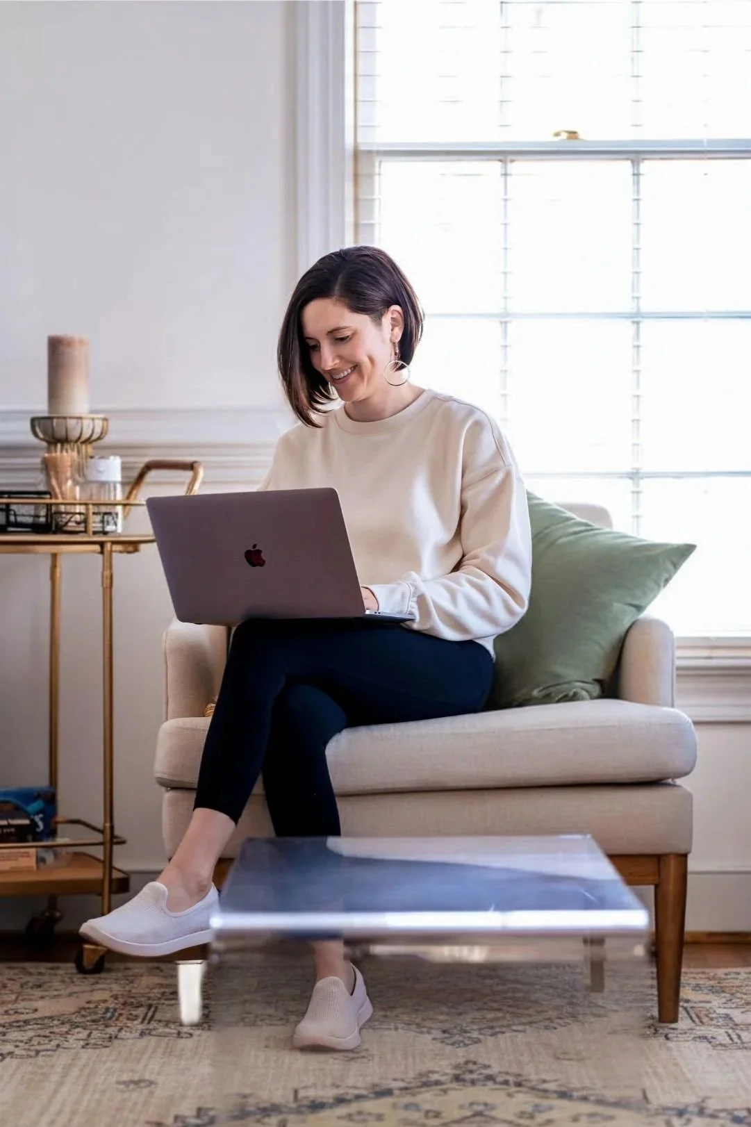 A woman sitting on a beige sofa with a green pillow, using a silver MacBook laptop, in a well-lit room with large windows, a side table with decorative items, and a glass coffee table in front.