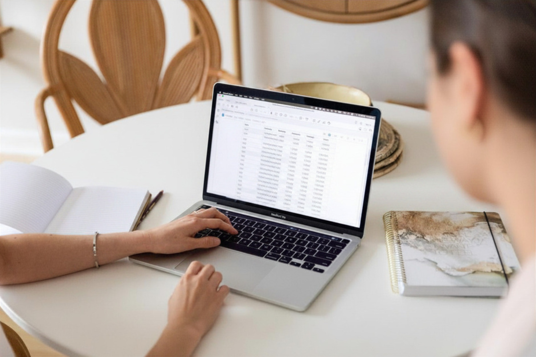 A person working on a laptop at a white table, with a spreadsheet on the screen, a notebook, and an open planner nearby. Another person is sitting beside them. In the background, there are rattan chairs and a decorative bowl.