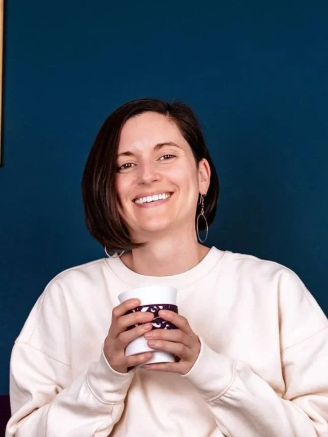 A young woman with short dark hair smiling while holding a paper cup with both hands against a plain dark blue background.