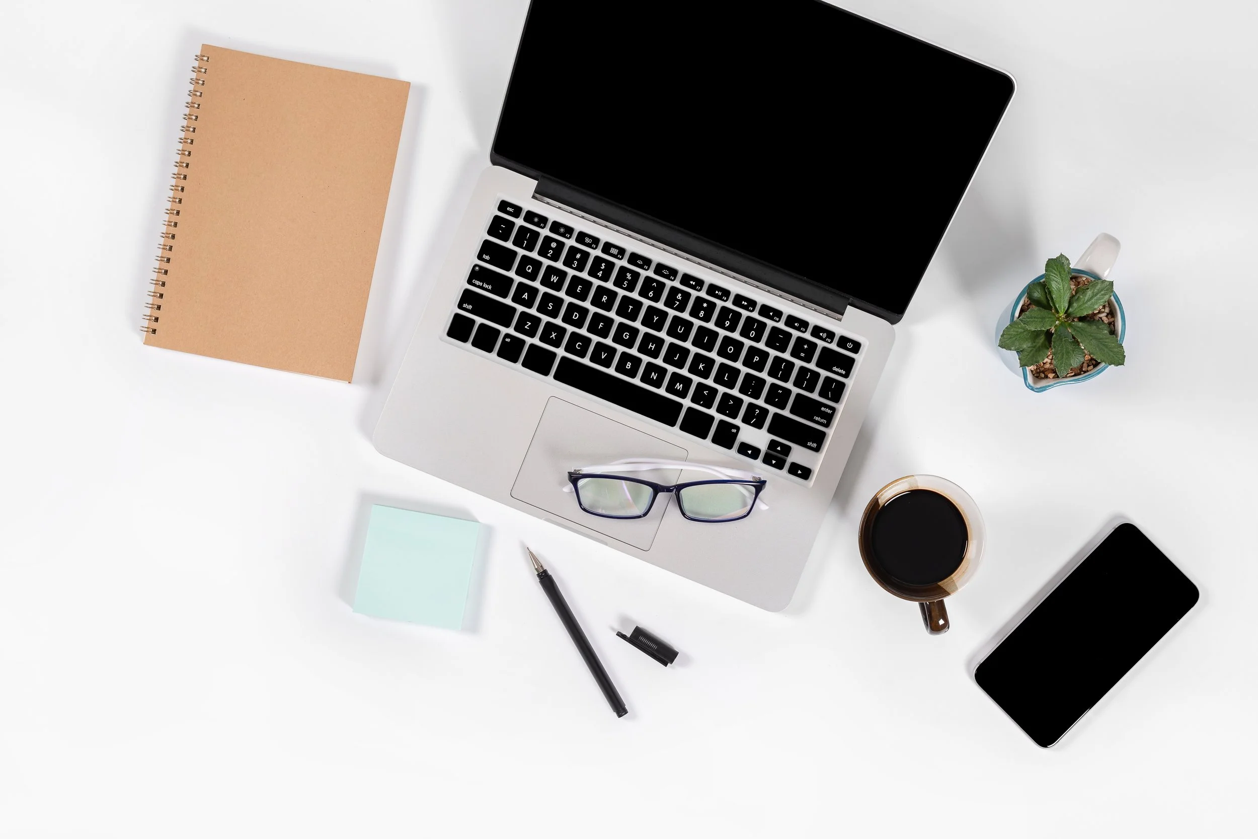 Top-down view of a workspace with a silver laptop, a smartphone, a pair of glasses, a notebook, sticky notes, a pen, a cup of coffee, and a small potted plant on a white surface.