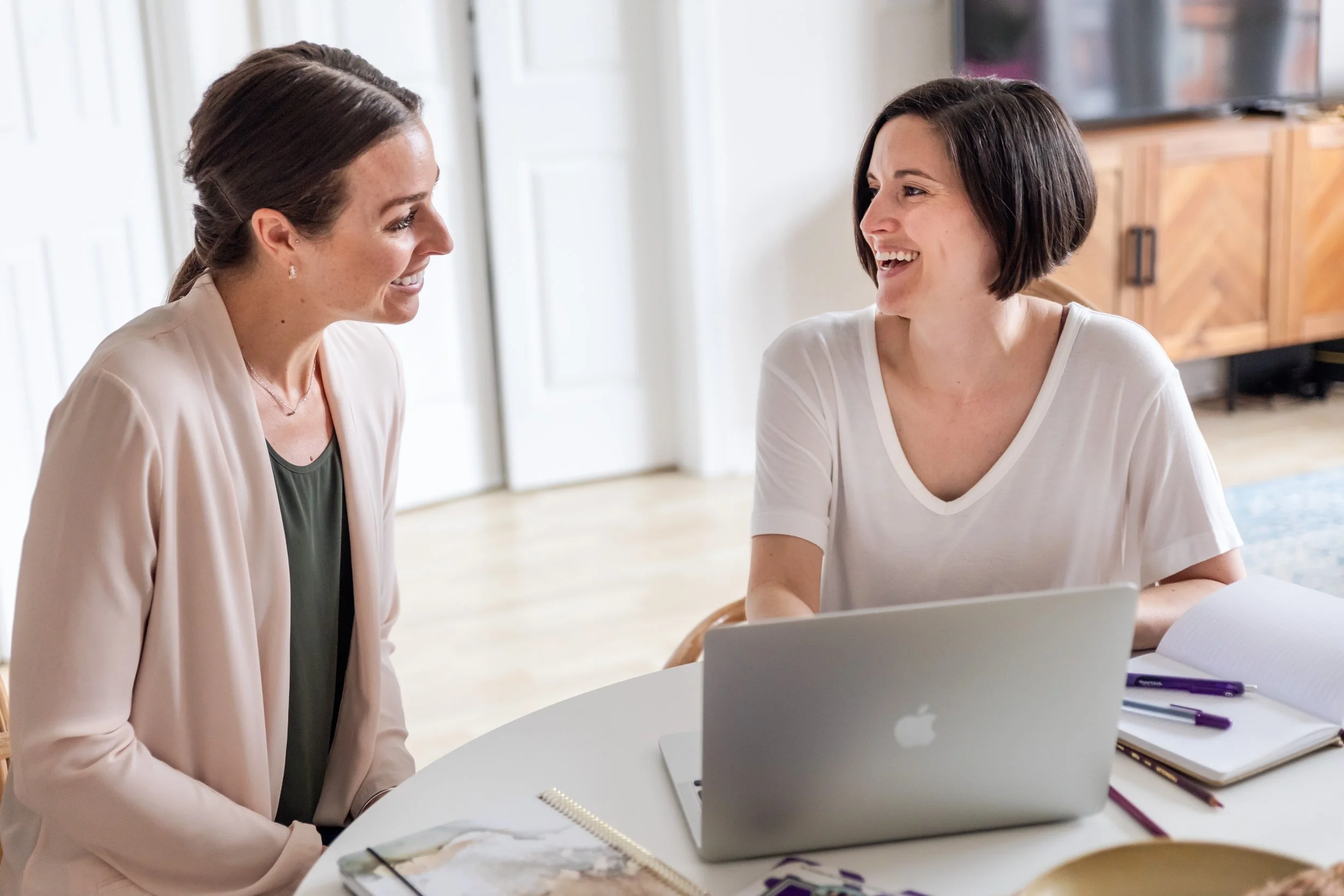 Two women smiling at each other while working on a laptop at a table with notebooks and pens.