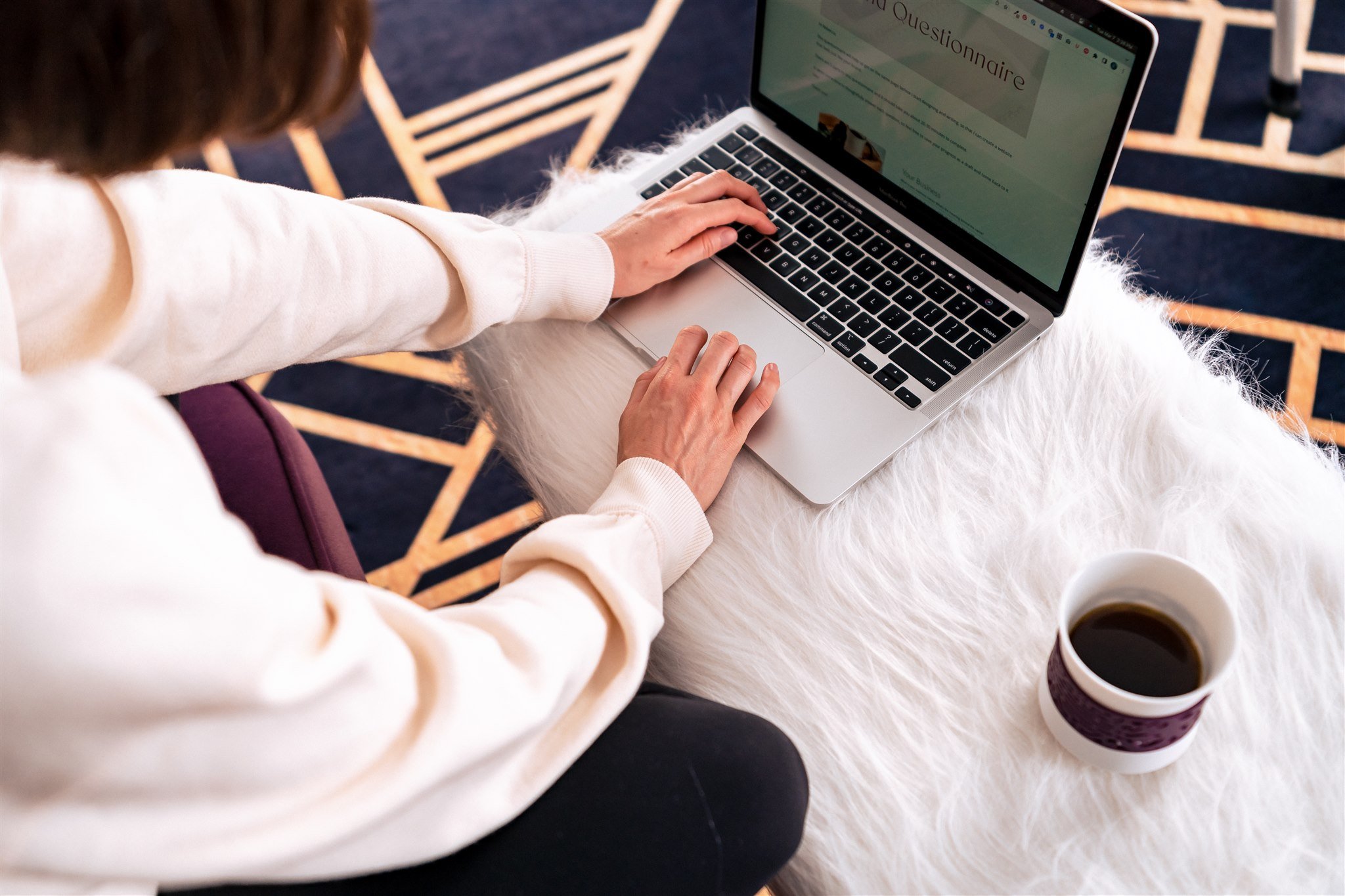 Looking over the shoulder of a woman working on a computer. The computer is sitting on a faux fur white ottoman and there is a cup of tea next to it.