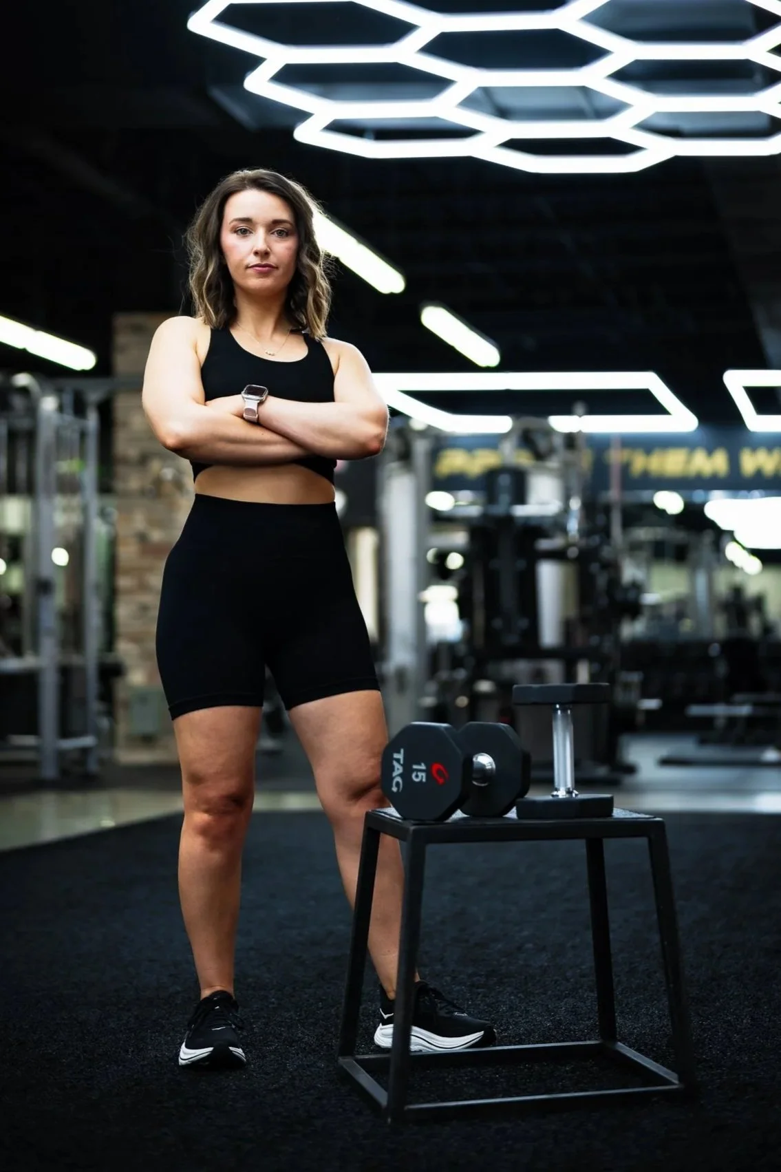 A woman standing with arms crossed in a gym, wearing black workout clothes, with workout equipment and modern lighting in the background.
