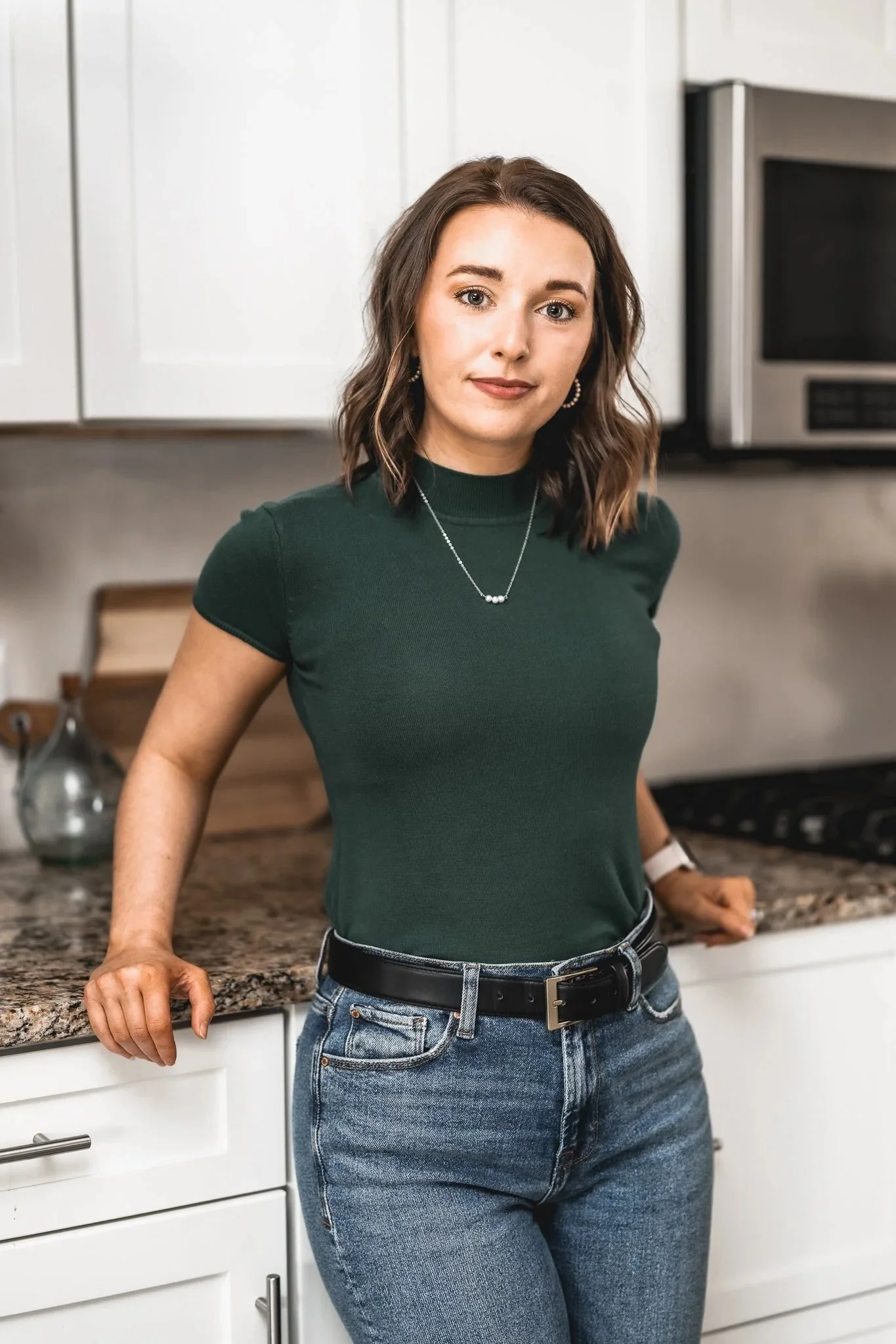 A woman with shoulder-length brown hair wearing a dark green top and blue jeans standing in a modern kitchen.