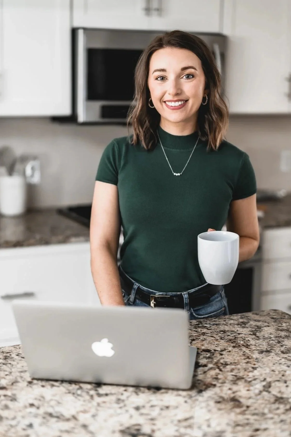 A woman with shoulder-length wavy hair, wearing a dark green t-shirt, jewelry, and a belt, smiling and holding a white mug in a kitchen with a granite countertop, white cabinets, and a microwave in the background.