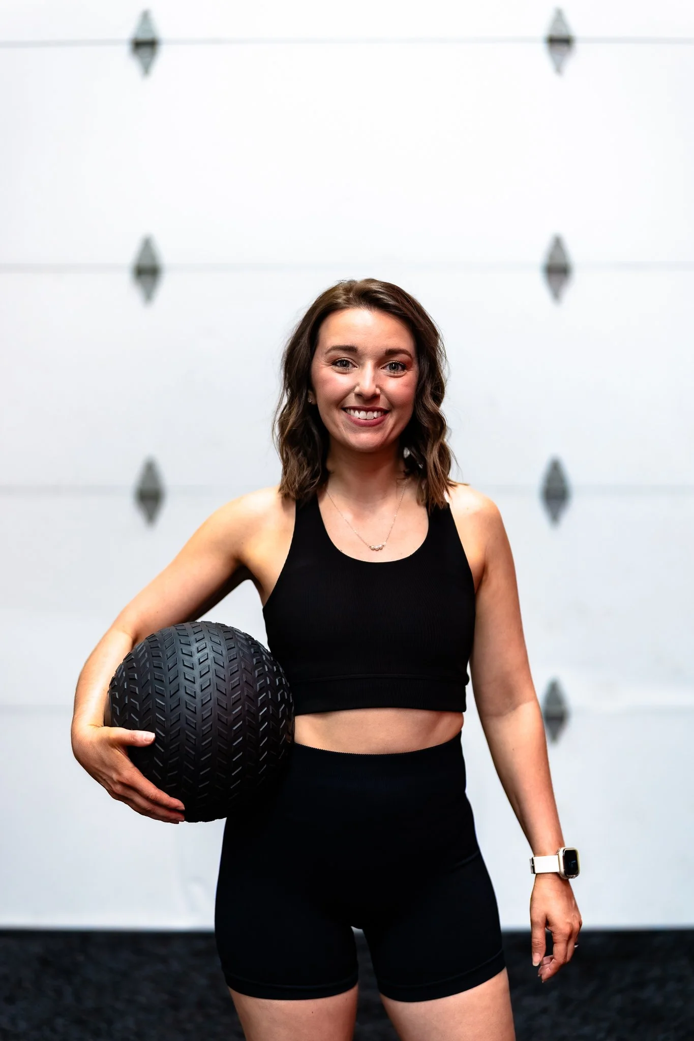 A young woman in athletic wear holding a black textured medicine ball, standing in front of a white garage door.