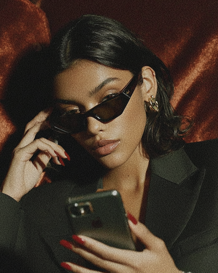 Woman with dark hair wearing sunglasses, earrings, and a black blazer, looking at smartphone against a dark textured background.
