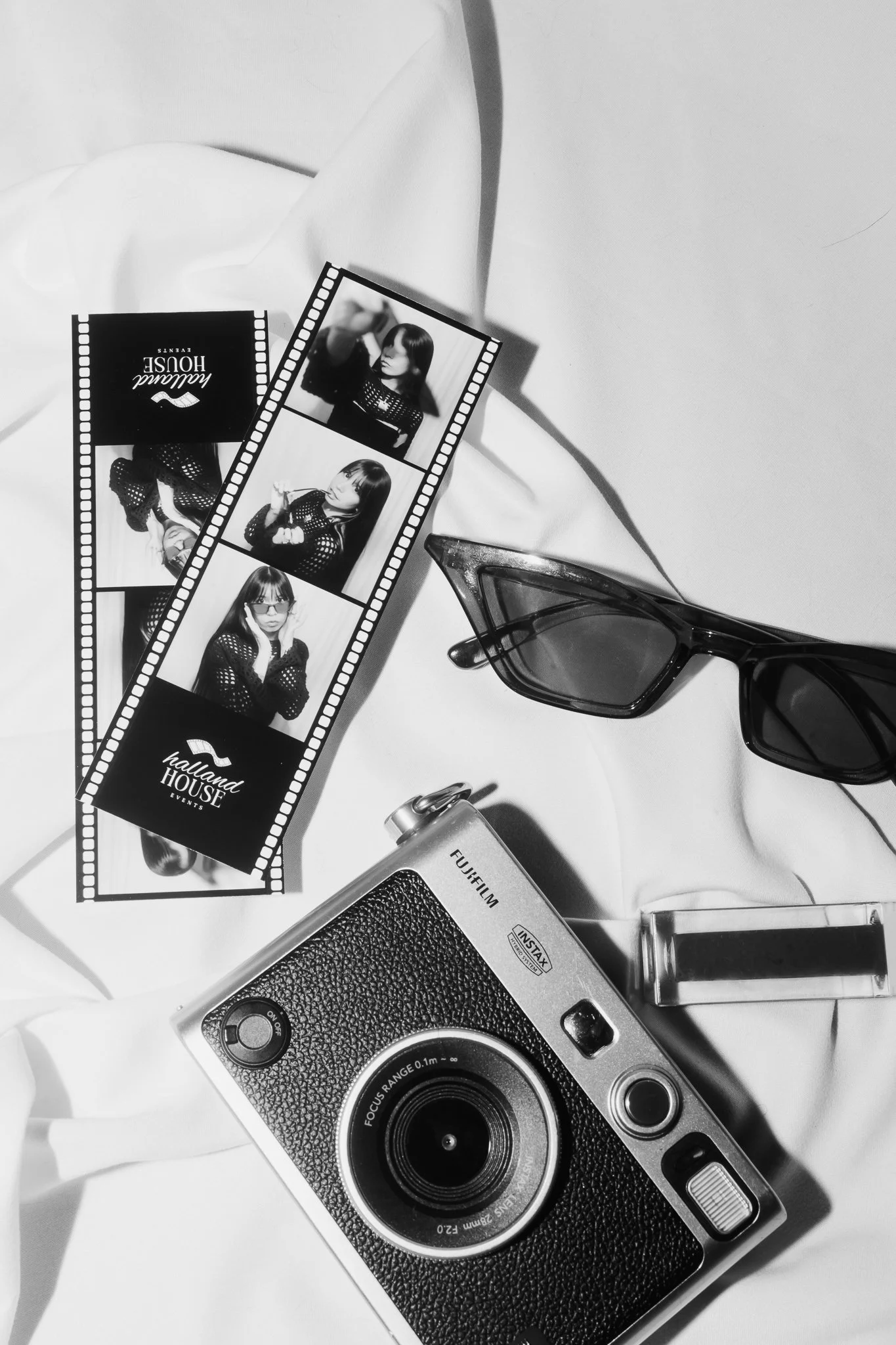A vintage film camera, sunglasses, a lipstick, and a strip of photo booth pictures on a white surface.