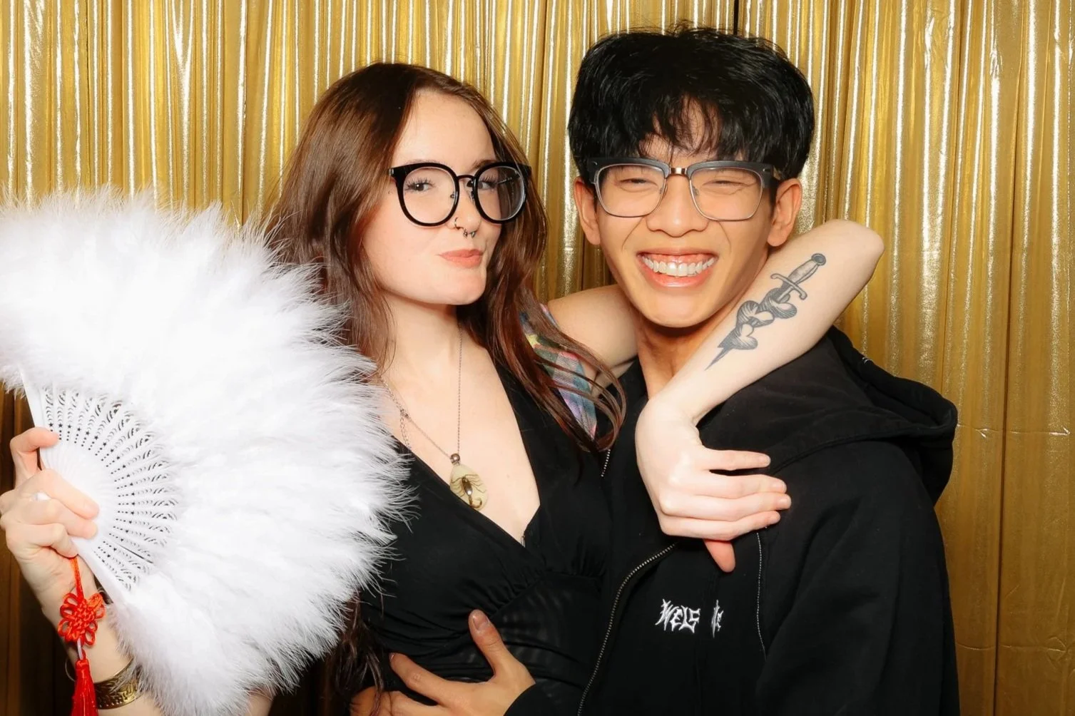 Two friends smiling and posing together in front of a gold curtain background, one holding a white feather fan.