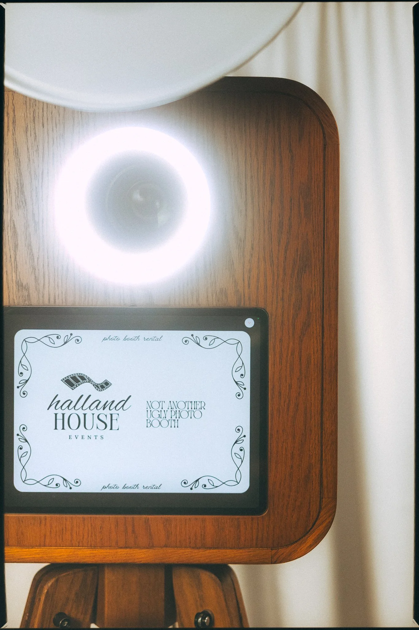 Social media or event flyer displayed on a tablet screen resting on a wooden table. The flyer reads "Halland House Events" with a filmstrip graphic, and mentions photo booth rental with the phrase "Not another ugly photo booth."