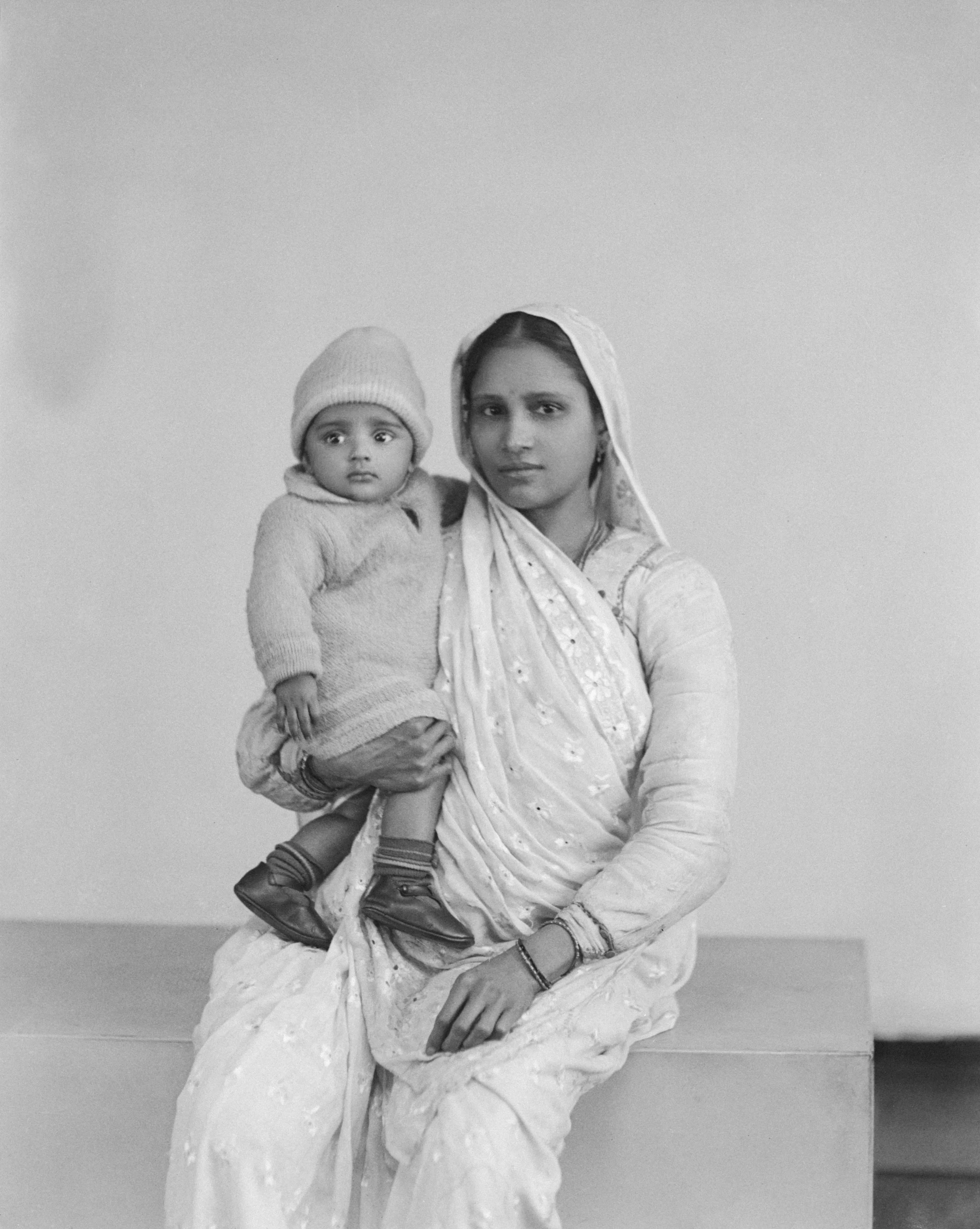 A woman in traditional Indian attire holding a small child dressed in a sweater, hat, and shoes, against a plain background.