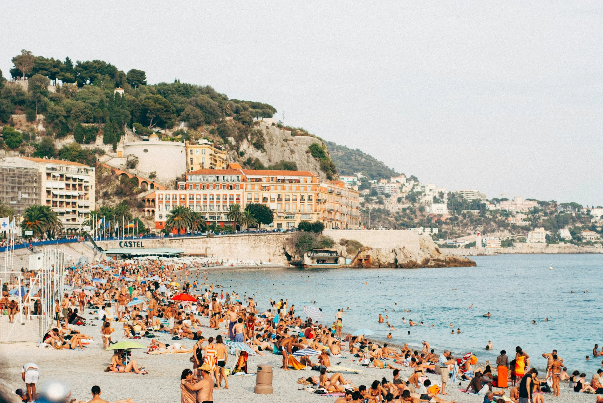 Crowded beach with many sunbathers, umbrellas, and swimmers by the shoreline, with colorful buildings and green hills in the background.