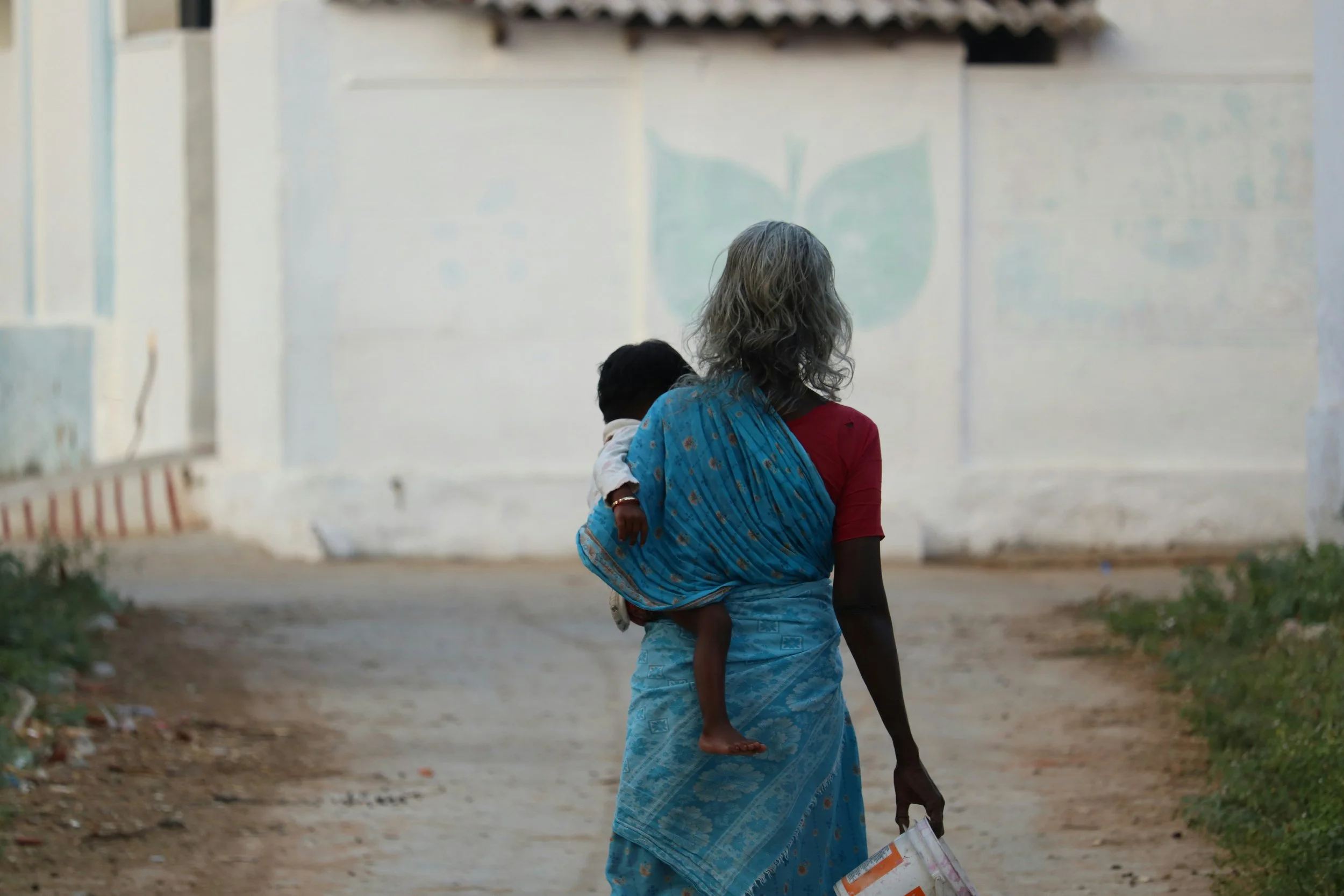 A woman walking outdoors carrying a young child on her back in a blue saree, holding a plastic bag, with a white wall in the background.