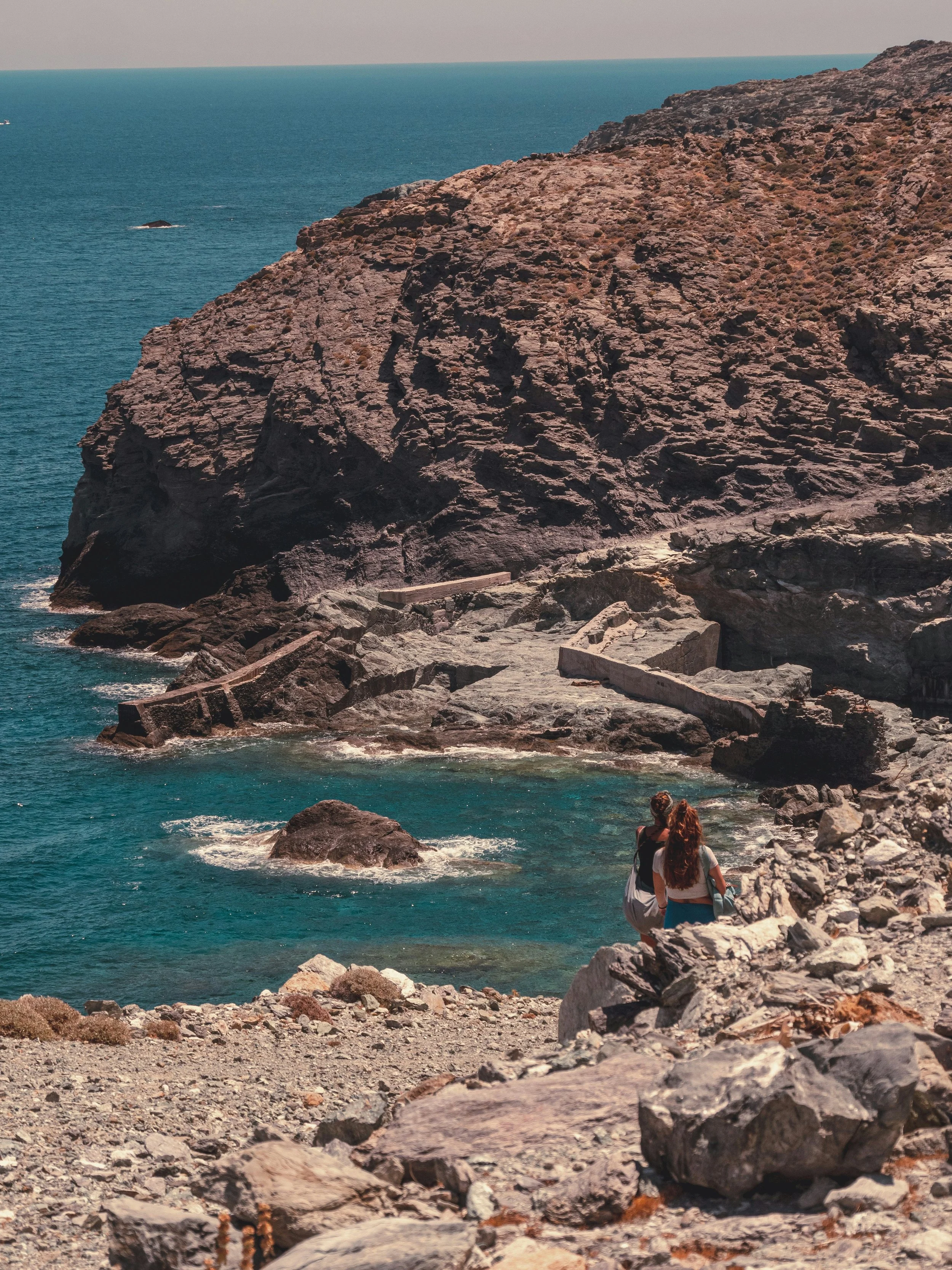 Two women sitting on rocks near the ocean, overlooking a rocky cliff and the horizon, with some boats in the distance.